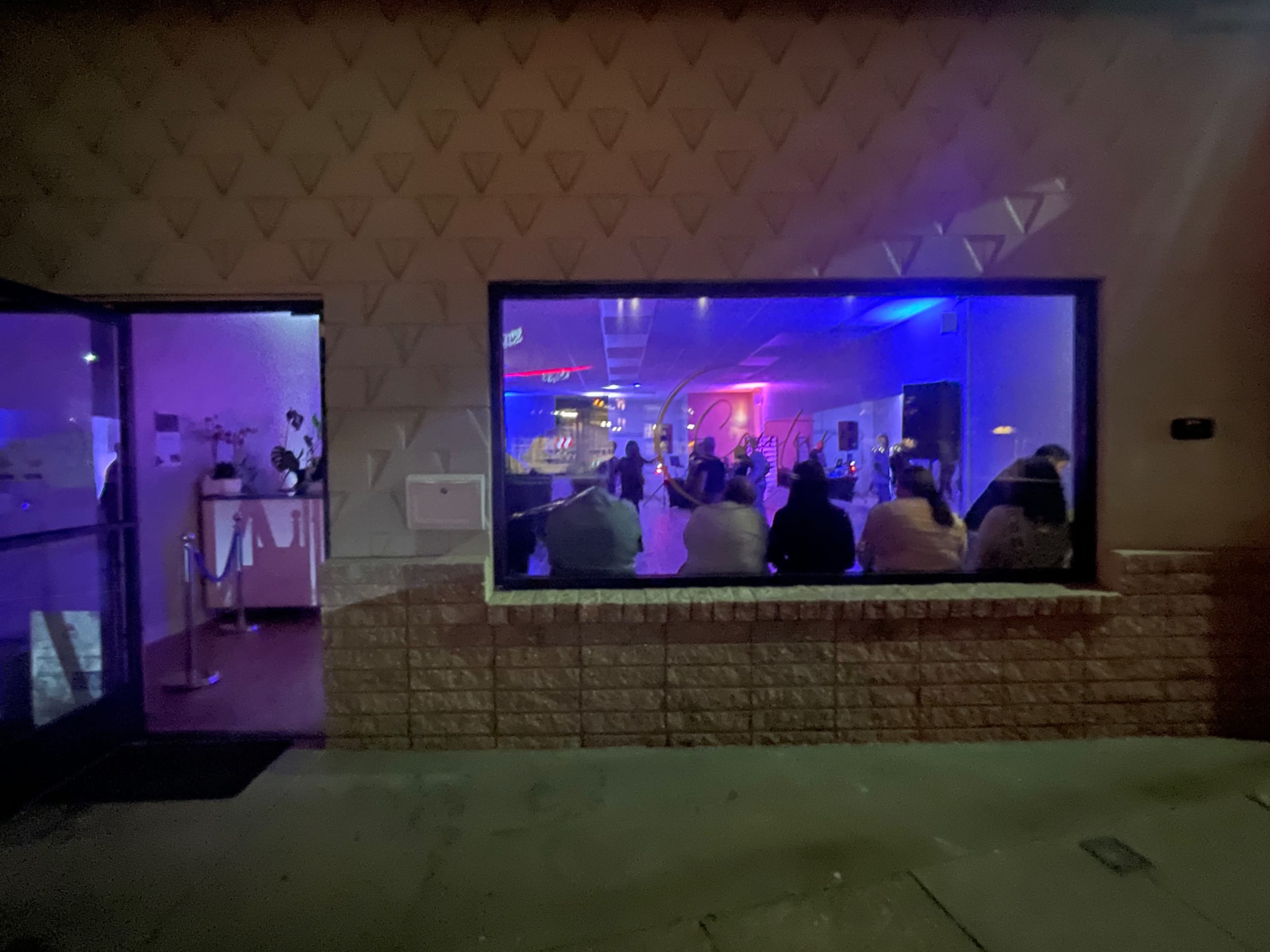 A group of people is seated in front of a brightly lit stage inside a venue, visible through a large window.