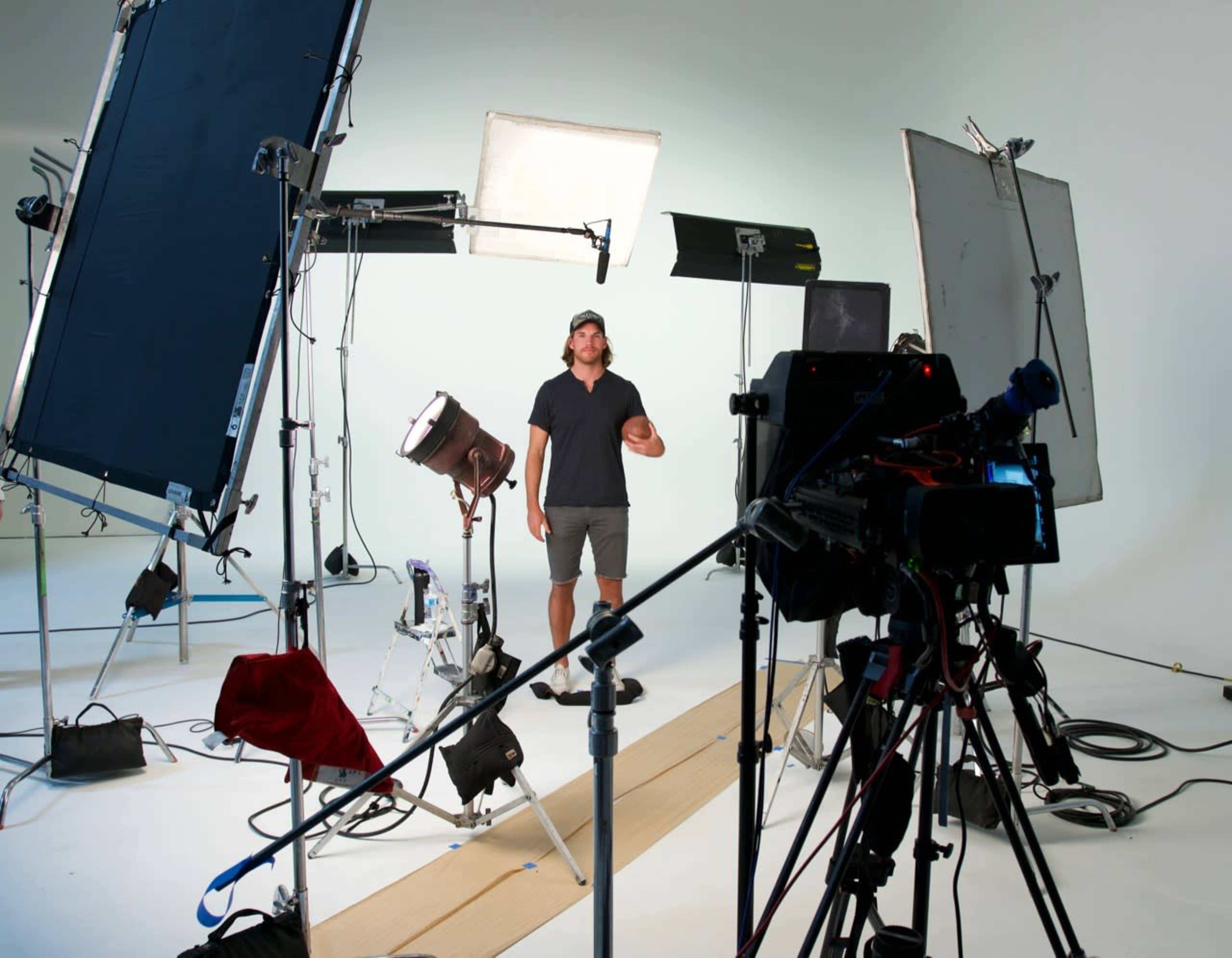 A man stands in a studio surrounded by lighting equipment and cameras on a white backdrop.