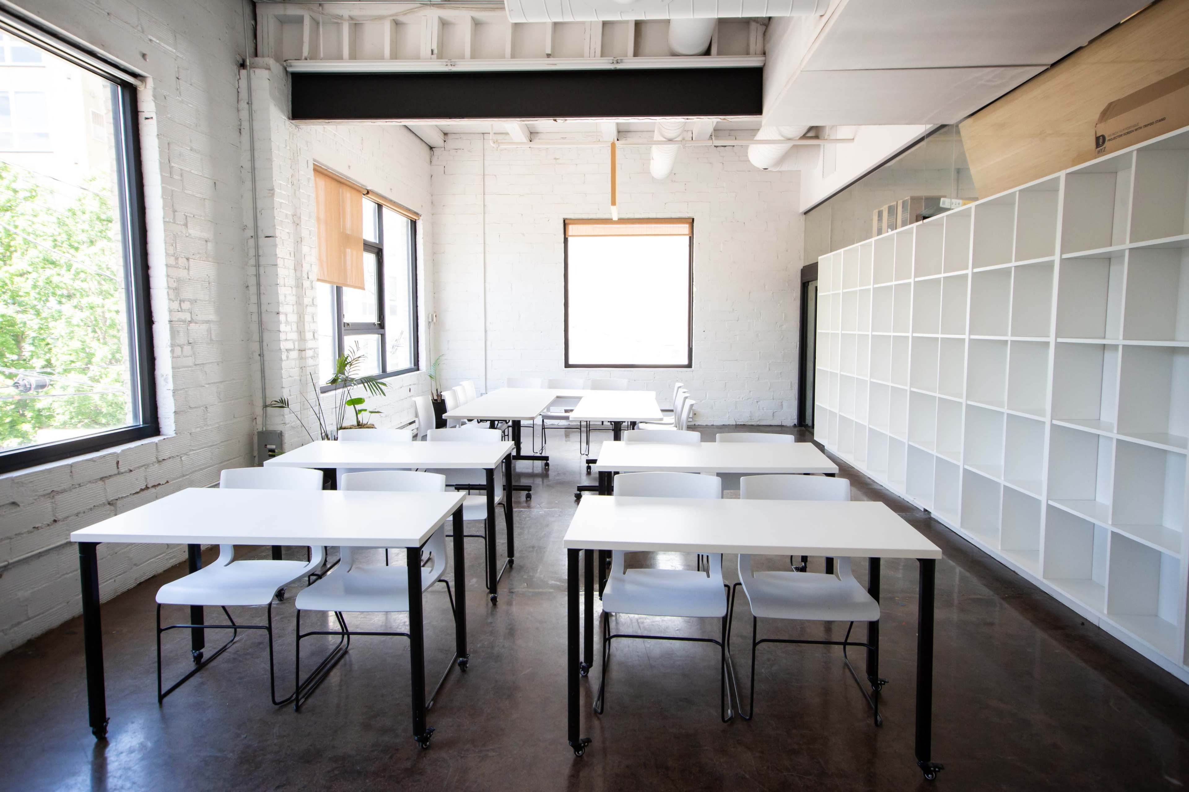 The image shows a bright, minimalist classroom with several white tables and chairs arranged in a clean, modern space.