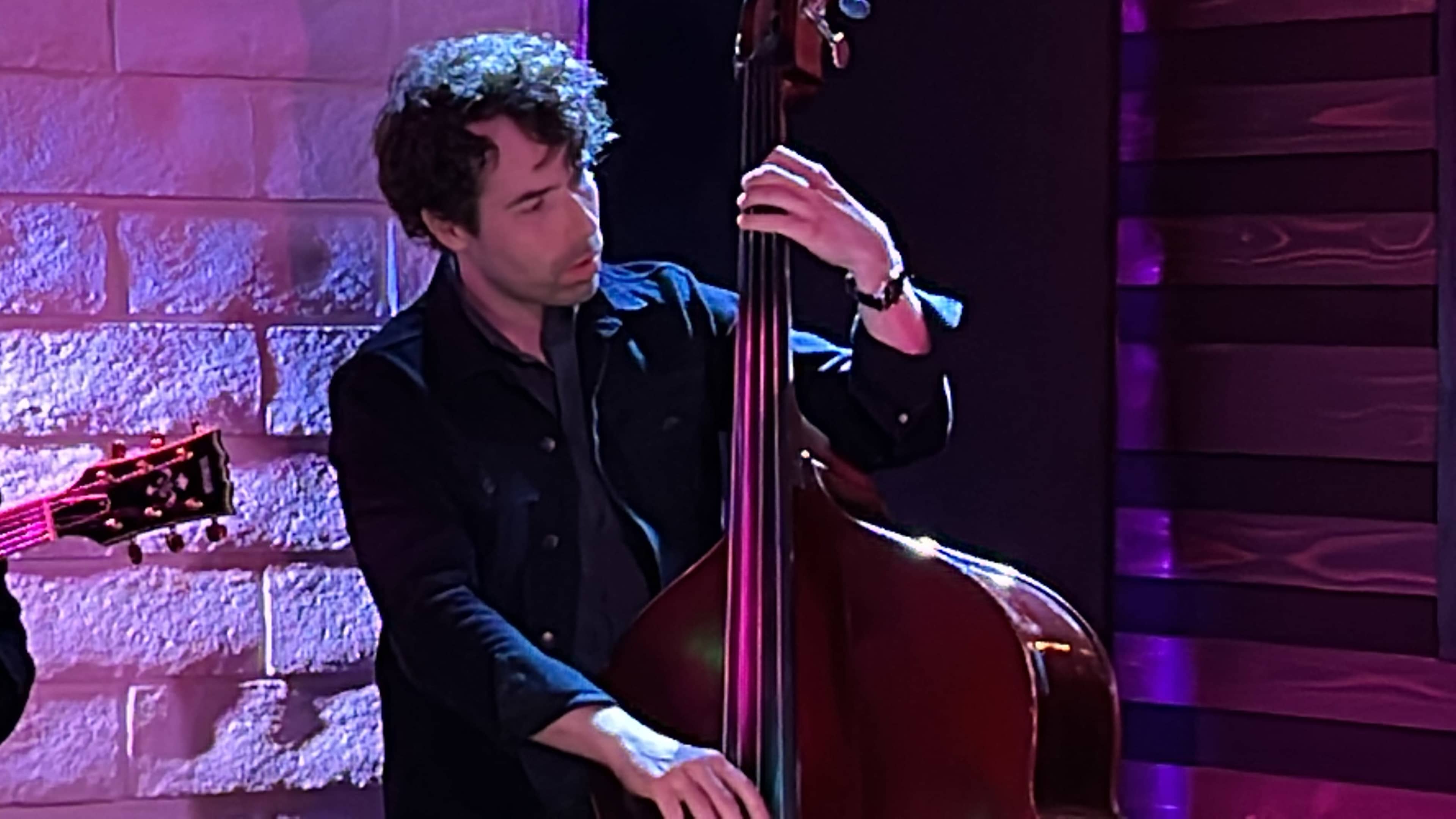 A musician plays a double bass on stage with colorful lighting in the background.