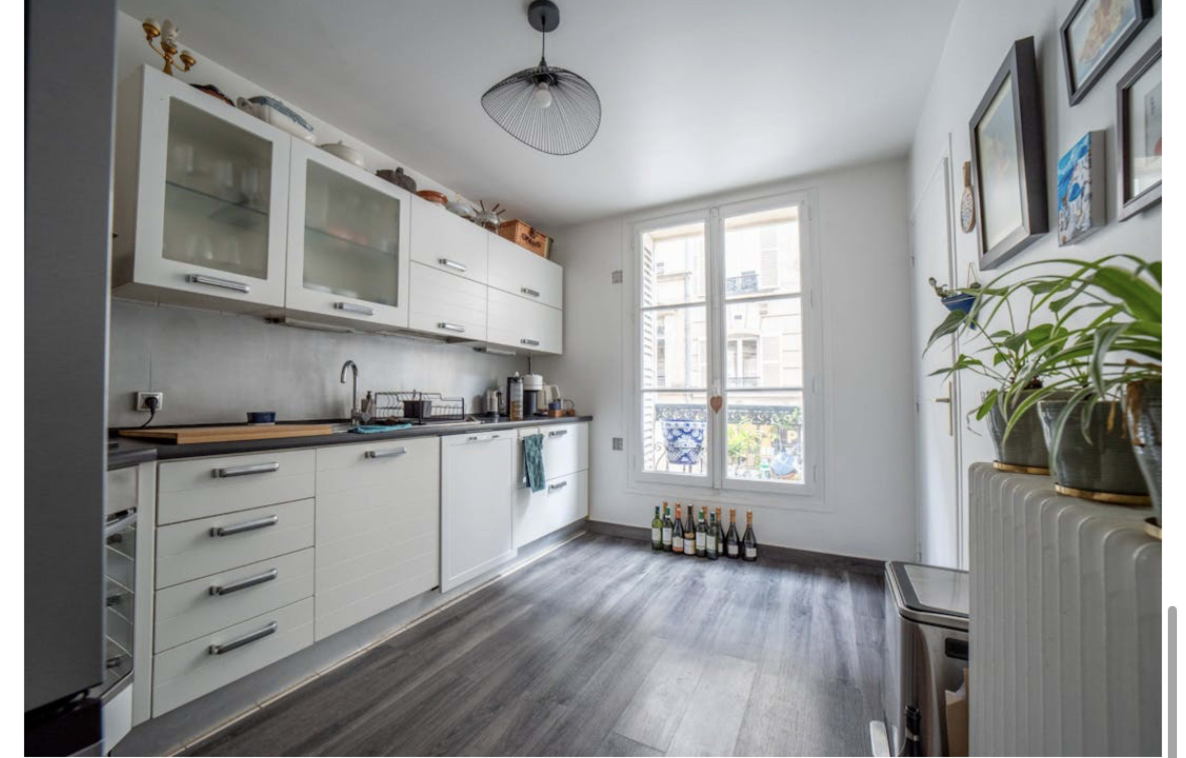 The image shows a modern kitchen with white cabinetry, a stainless steel sink, and a window overlooking a courtyard, along with a collection of wine bottles neatly arranged on the floor.