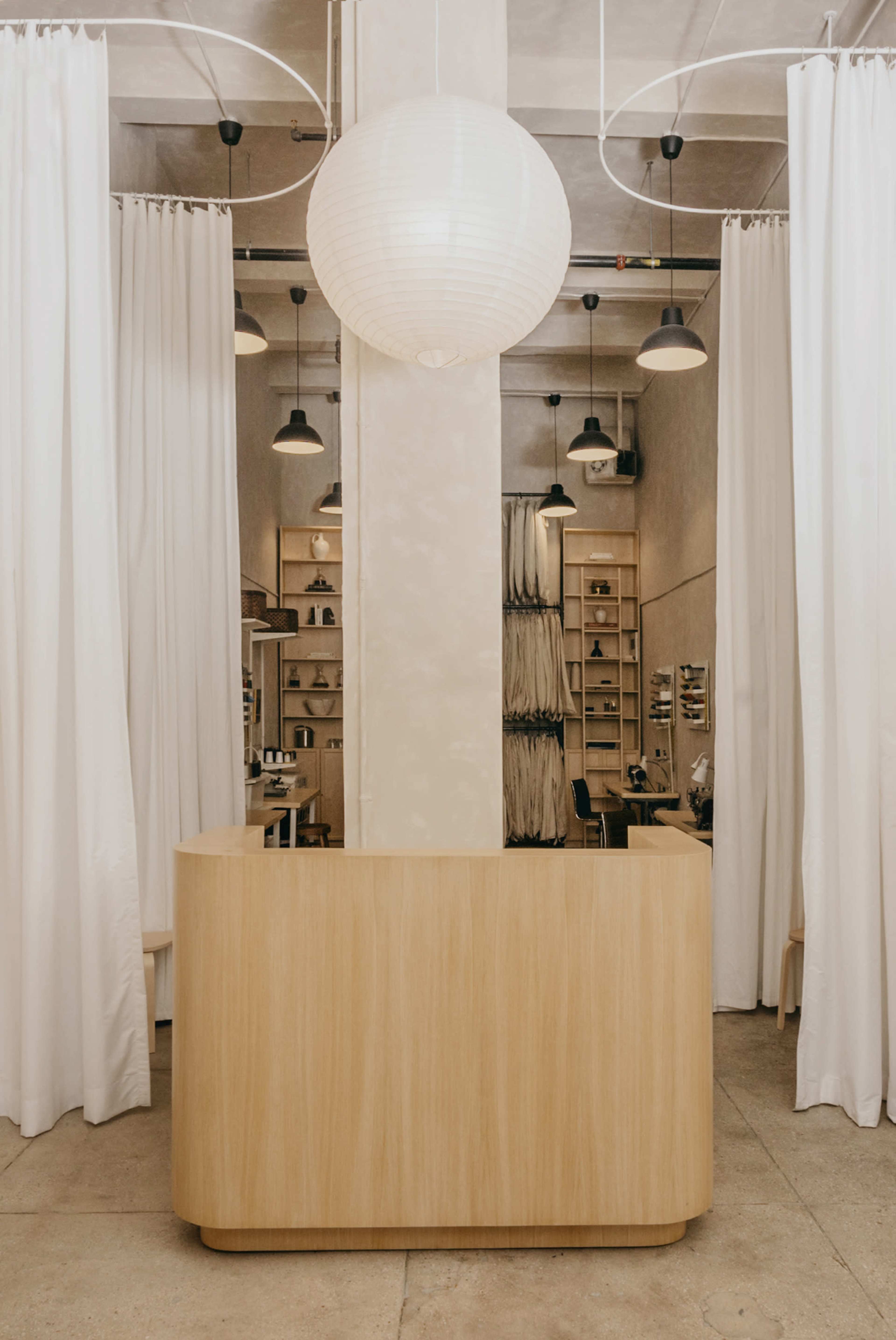 A wooden reception desk stands in a minimalist space with white curtains and a large paper lantern overhead.
