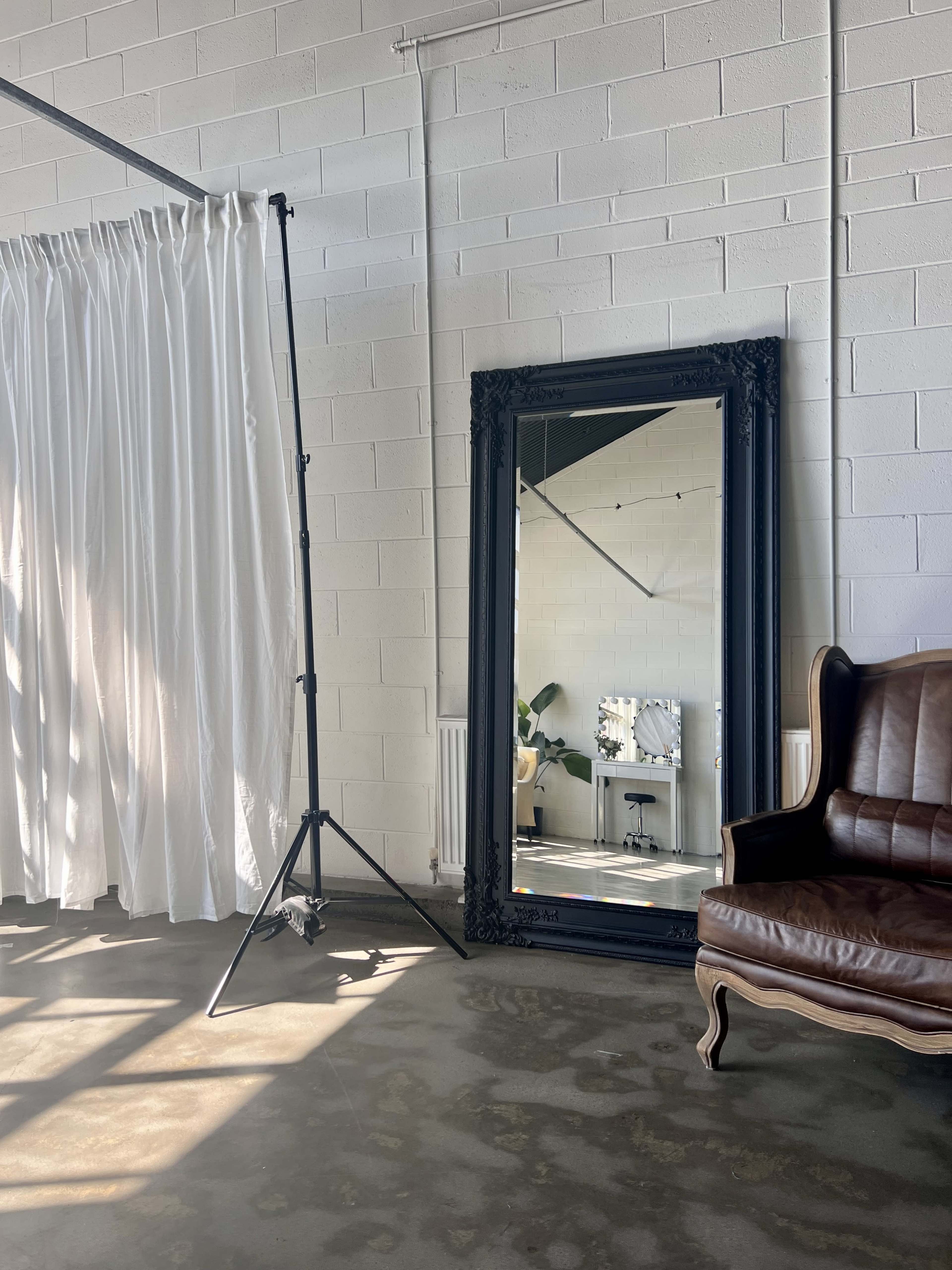 The image shows a large black ornate mirror reflecting a well-lit interior space with a curtain, a chair, and a plant.