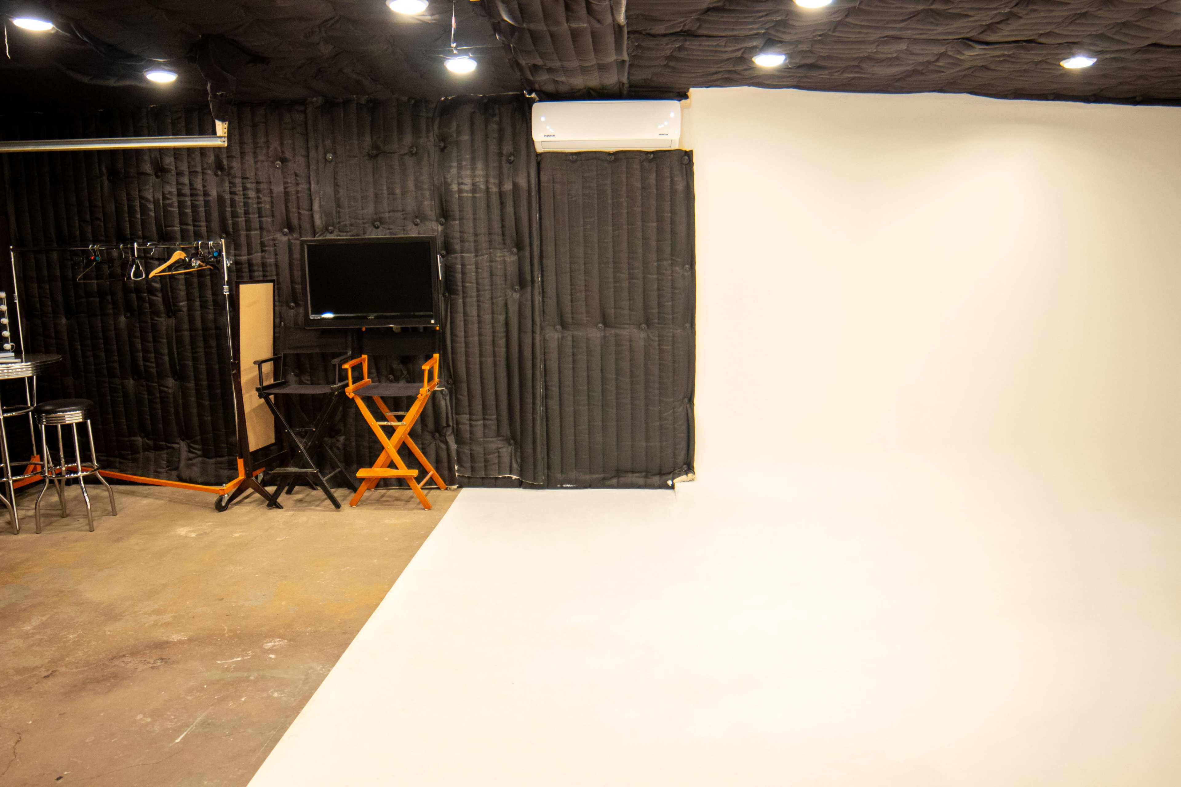 The image shows a photo studio with a white backdrop, a TV on a stand, a director's chair, and black soundproofing panels on the walls.