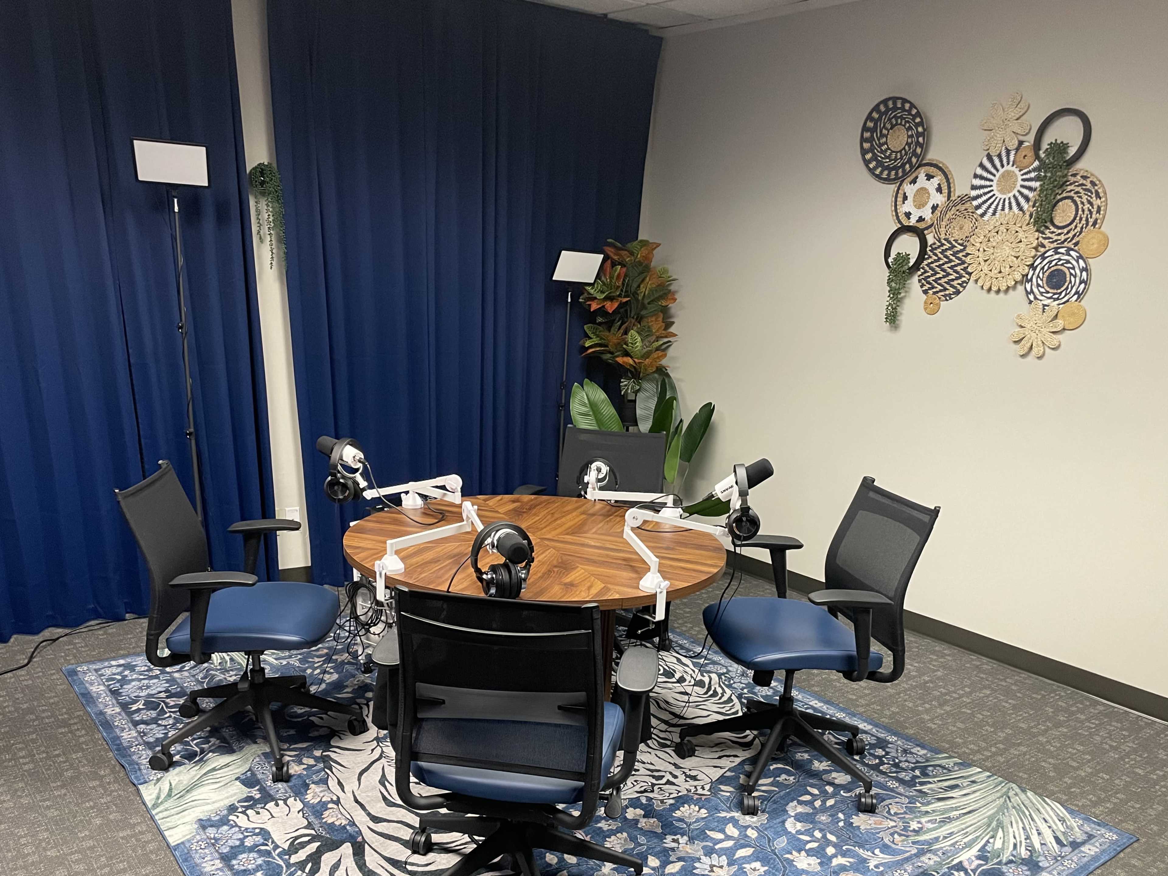 The image shows a small recording studio with a round table surrounded by four chairs, equipped with microphones and decorative elements on the walls.