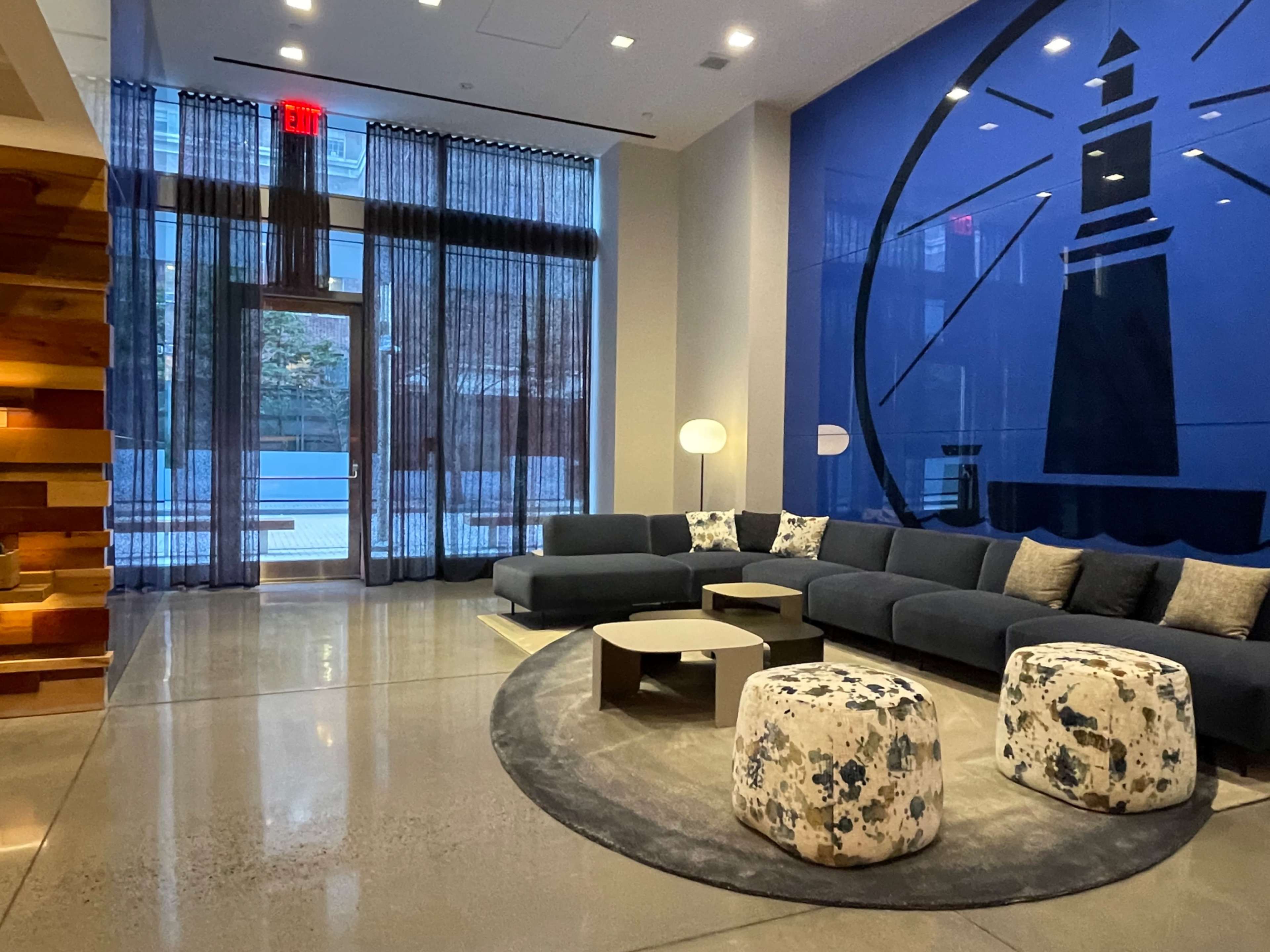 The image shows a modern lounge area featuring a large sectional sofa, round coffee tables, and decorative poufs against a backdrop of a blue wall with a lighthouse design.