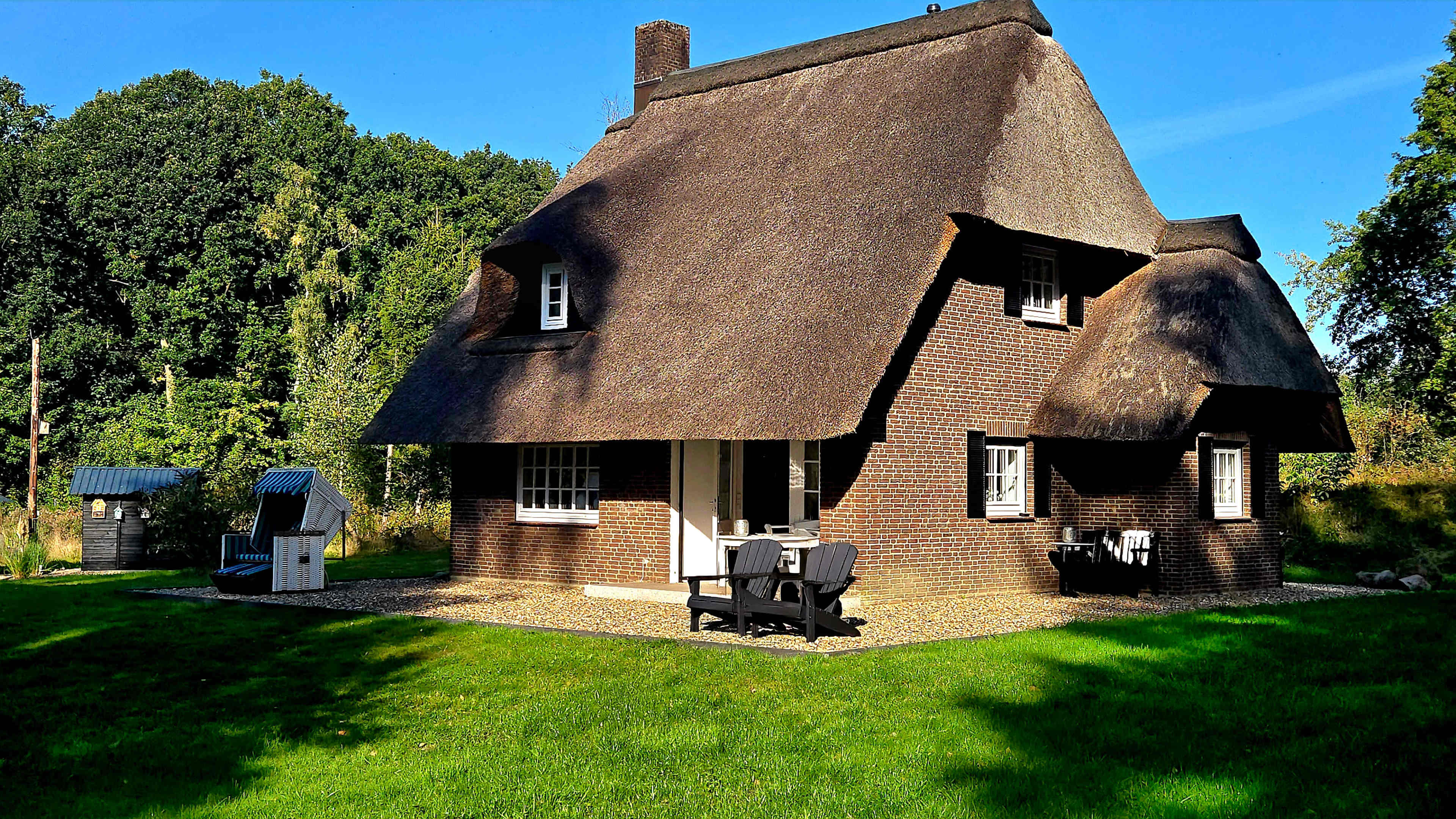 A thatched-roof house with brick walls and a grassy yard is surrounded by trees and features outdoor seating.