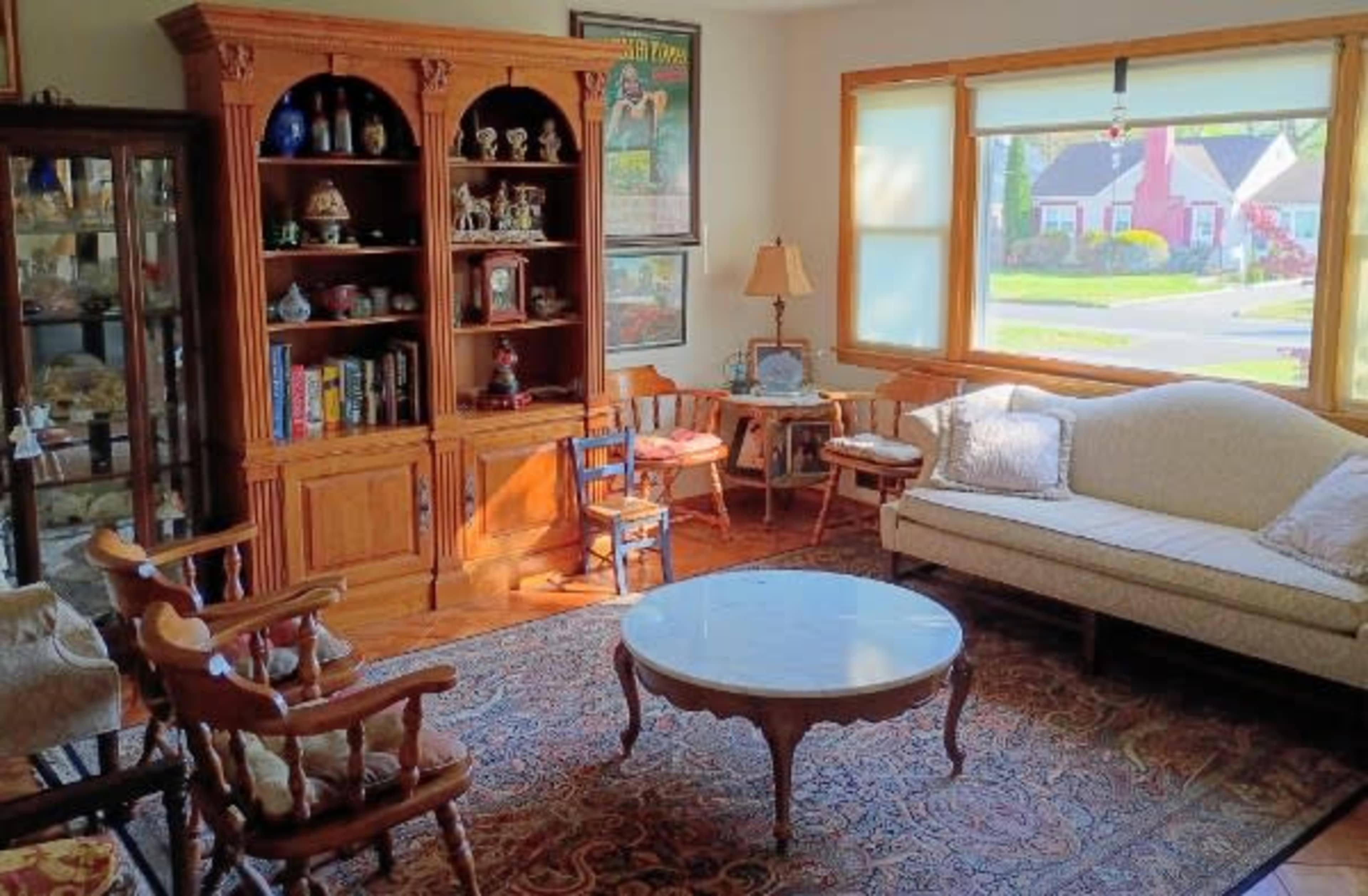 The room features a wooden bookshelf, a light-colored sofa, a round coffee table, and various decorative items arranged around a window that overlooks a neighborhood.