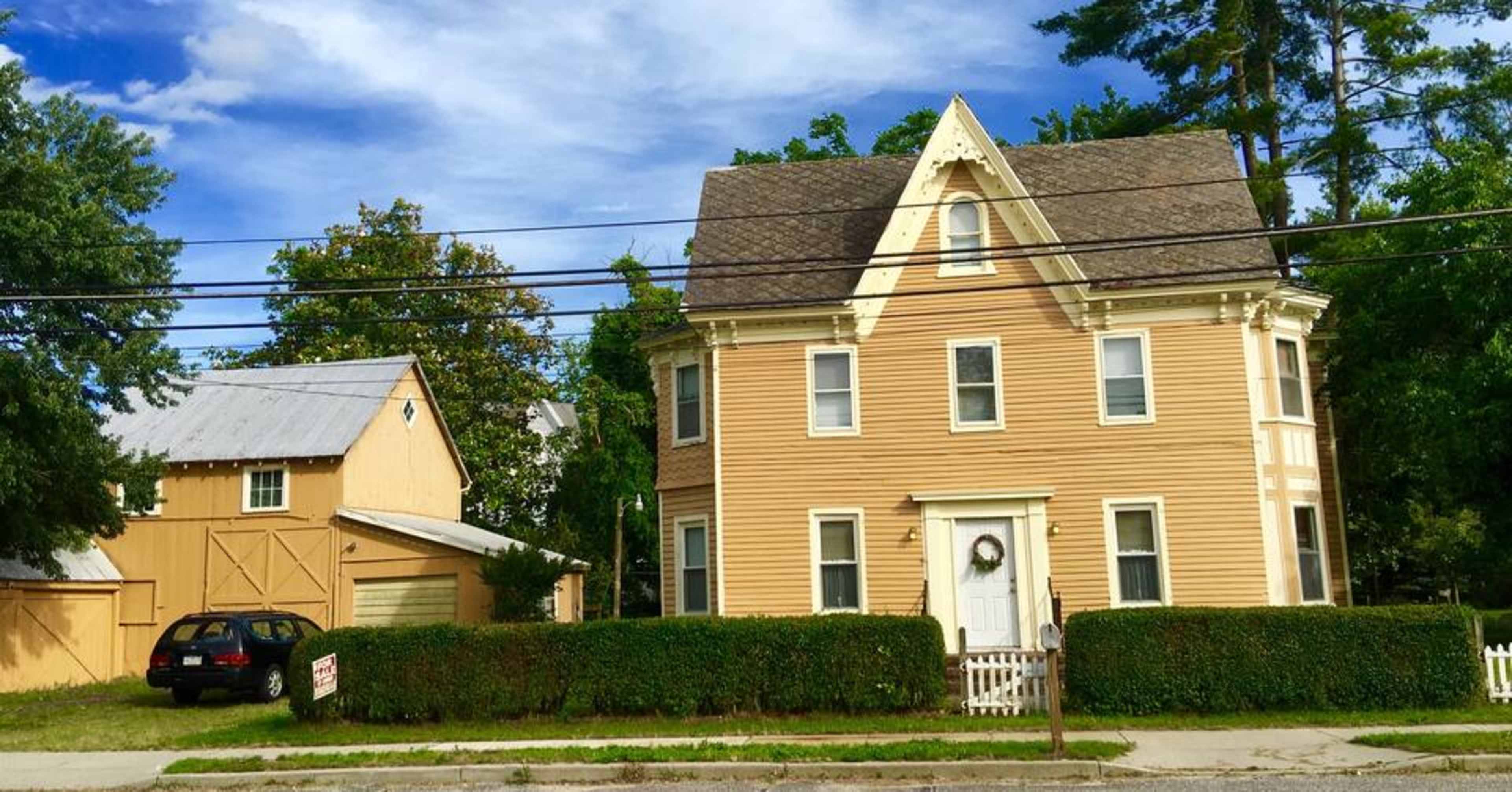 A two-story yellow house with a gabled roof next to a smaller yellow barn, surrounded by a neatly trimmed hedge and lined by power lines.