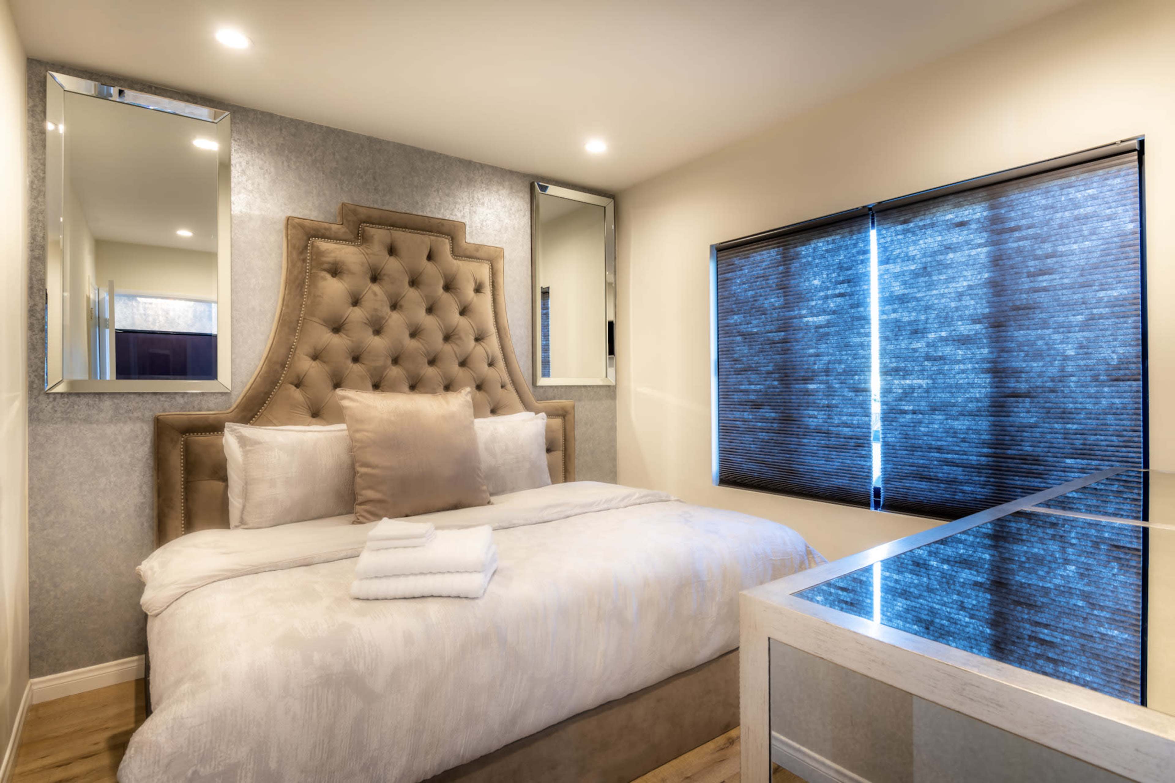 A modern bedroom featuring a tufted headboard, a light-colored bedding set, mirrored wall accents, and a window covered with dark blinds.