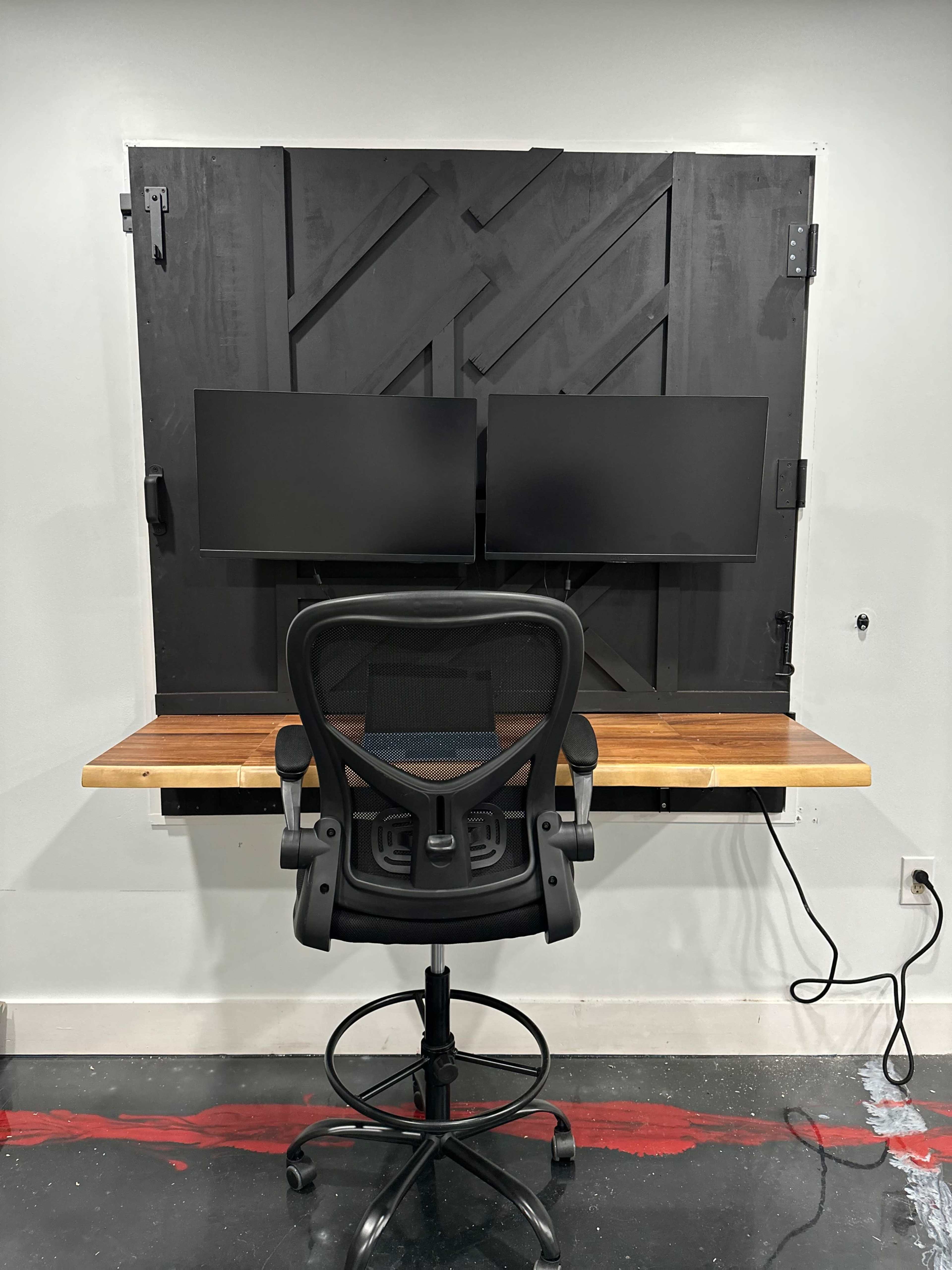 The image shows a minimalistic office setup featuring a wooden desk mounted on a wall, with two monitors above it and a black ergonomic chair in front.