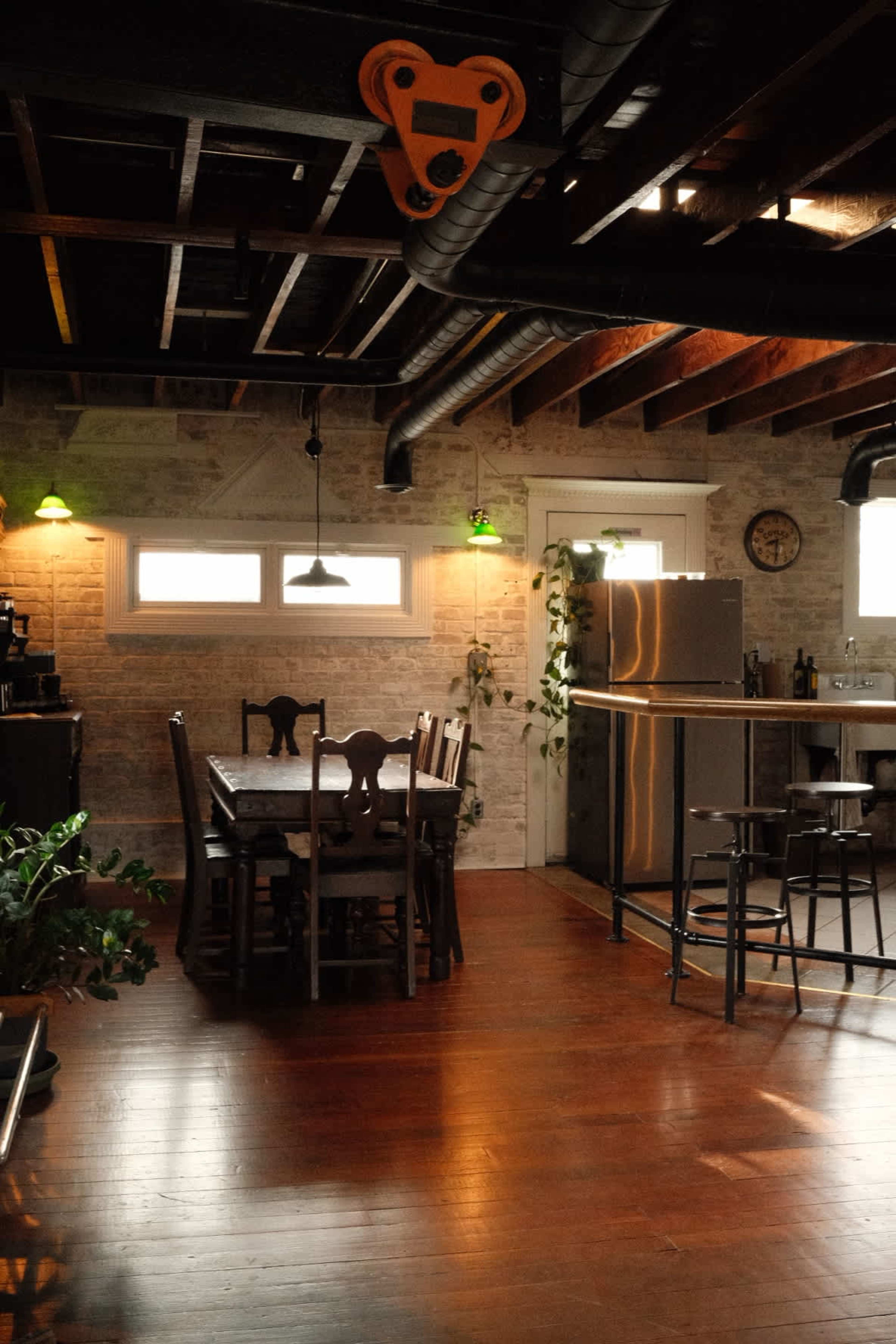 The image shows a modern kitchen and dining area with a wooden dining table, metal bar stools, and exposed beams in the ceiling.