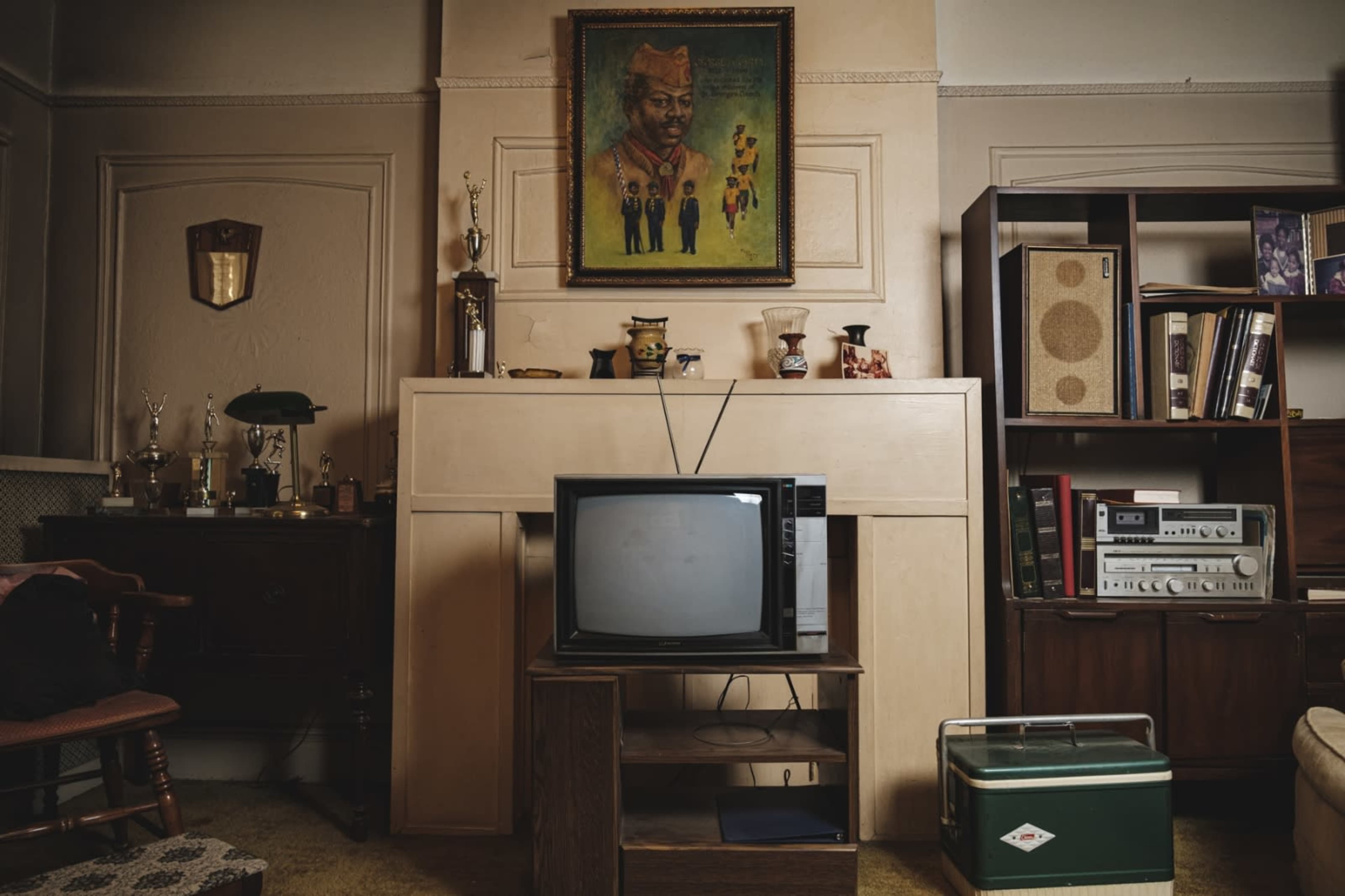 A retro living room with a vintage television set on a wooden stand, framed by a large painting and surrounded by various trophies and bookshelves.