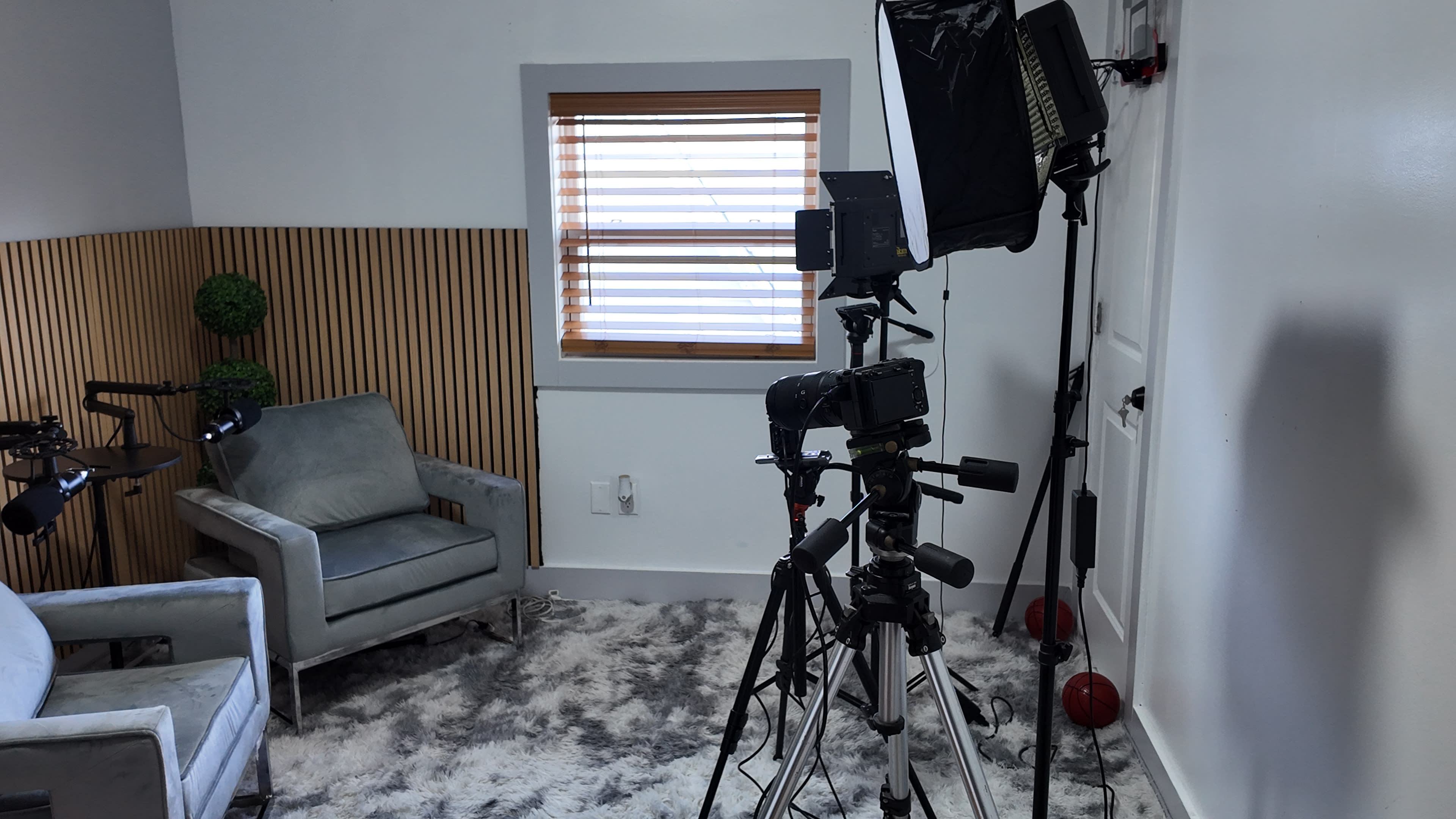 A well-lit room featuring a seated area with a light gray chair, a large window with blinds, and several tripods with lighting and camera equipment.