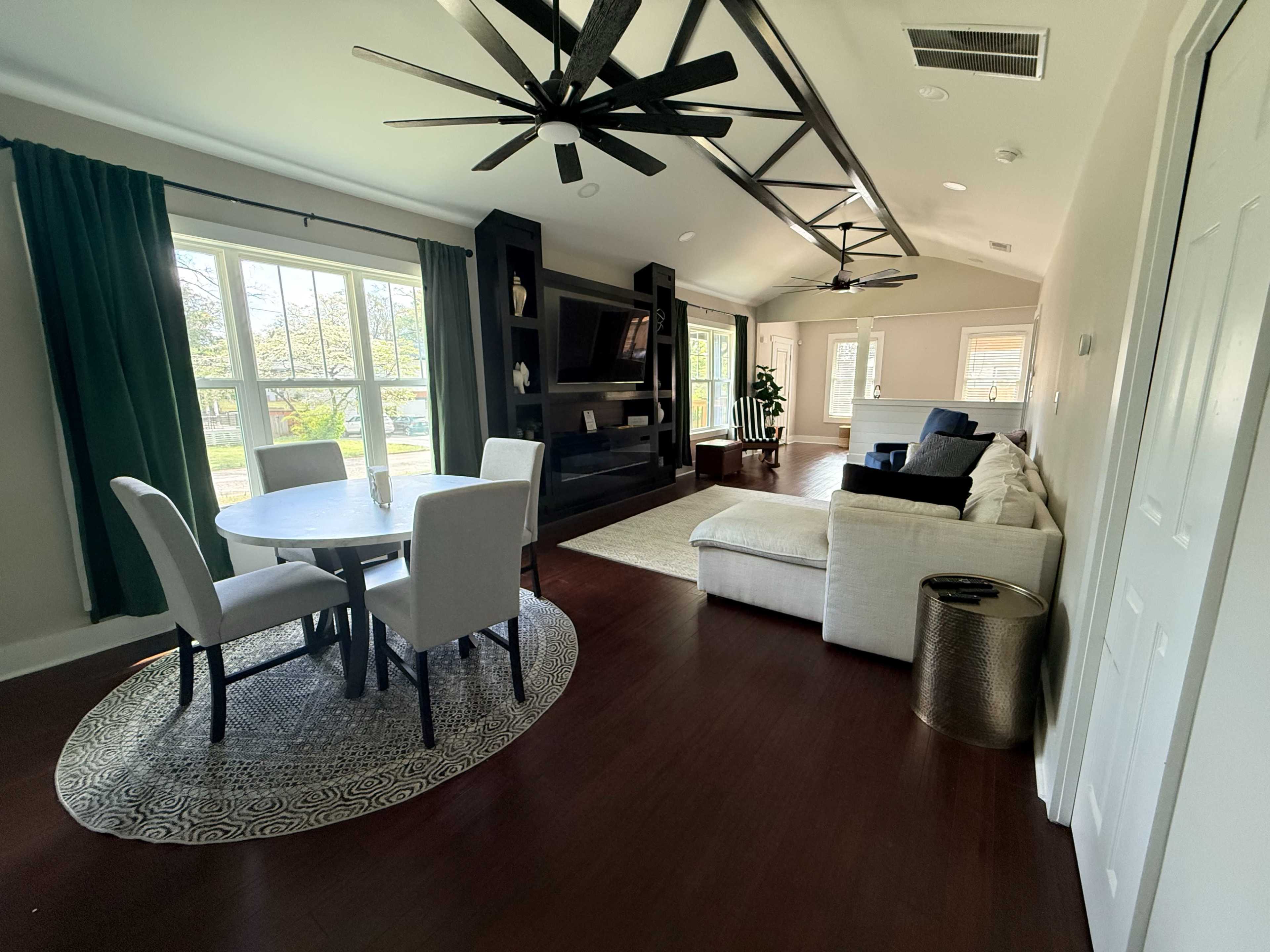 The image shows a spacious living room with a round dining table, a white sectional sofa, a large black entertainment unit, and large windows allowing natural light.