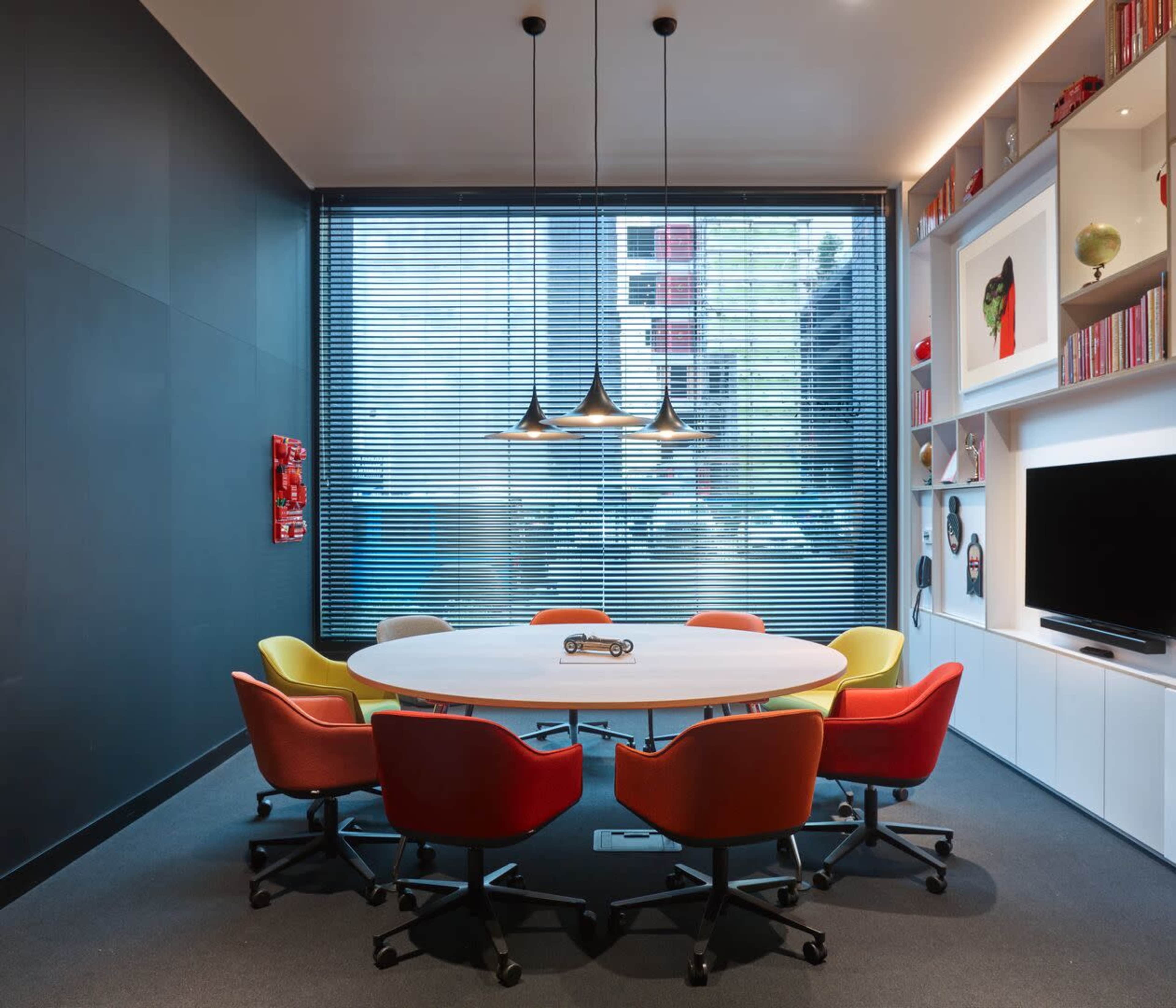A brightly lit conference room features a large oval table surrounded by eight colorful chairs, with a window displaying an urban view and shelves filled with decorative items and books.