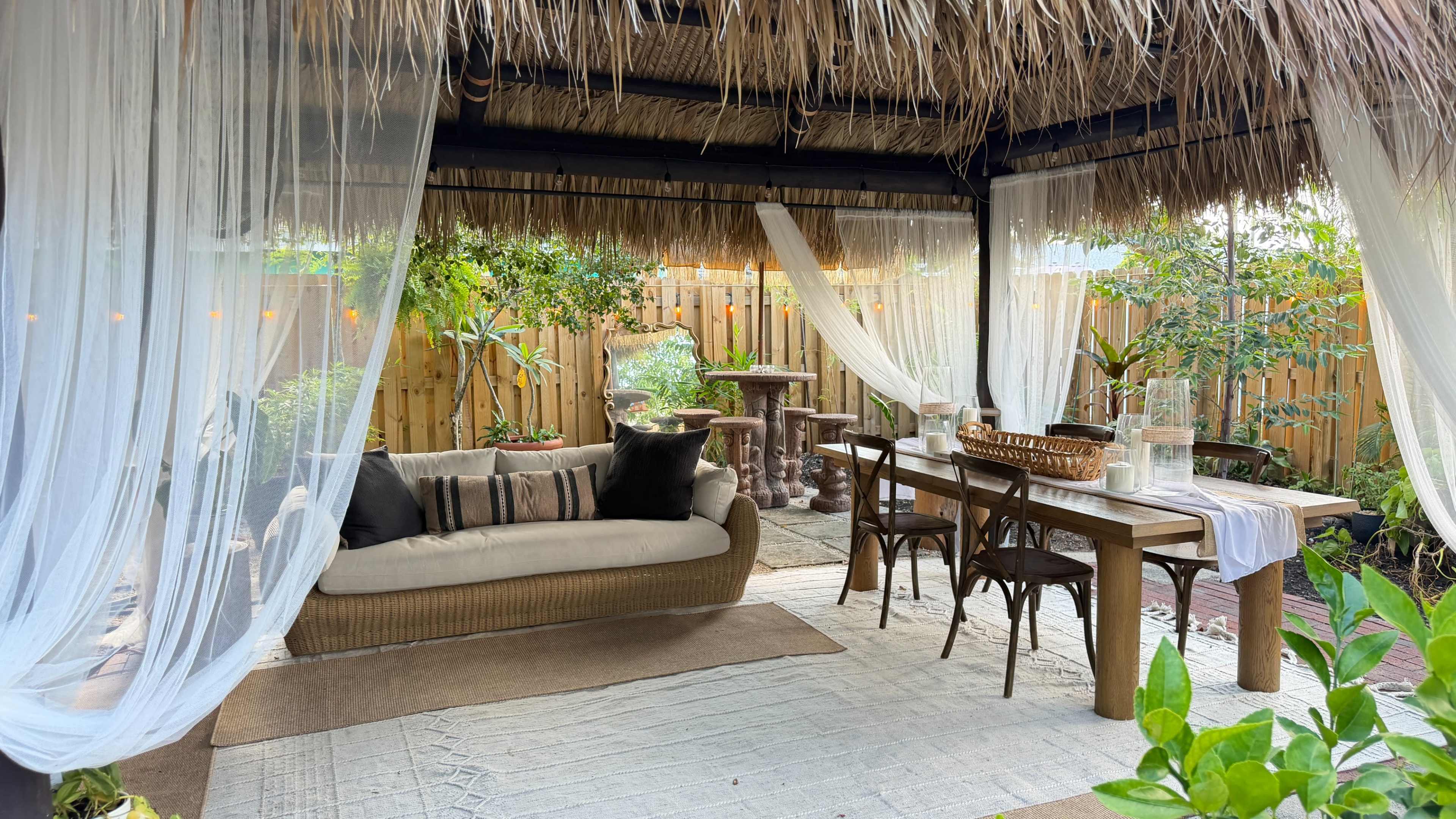 The image shows a shaded outdoor lounge area with a sofa, wooden table, and chairs, surrounded by plants and draped fabric.