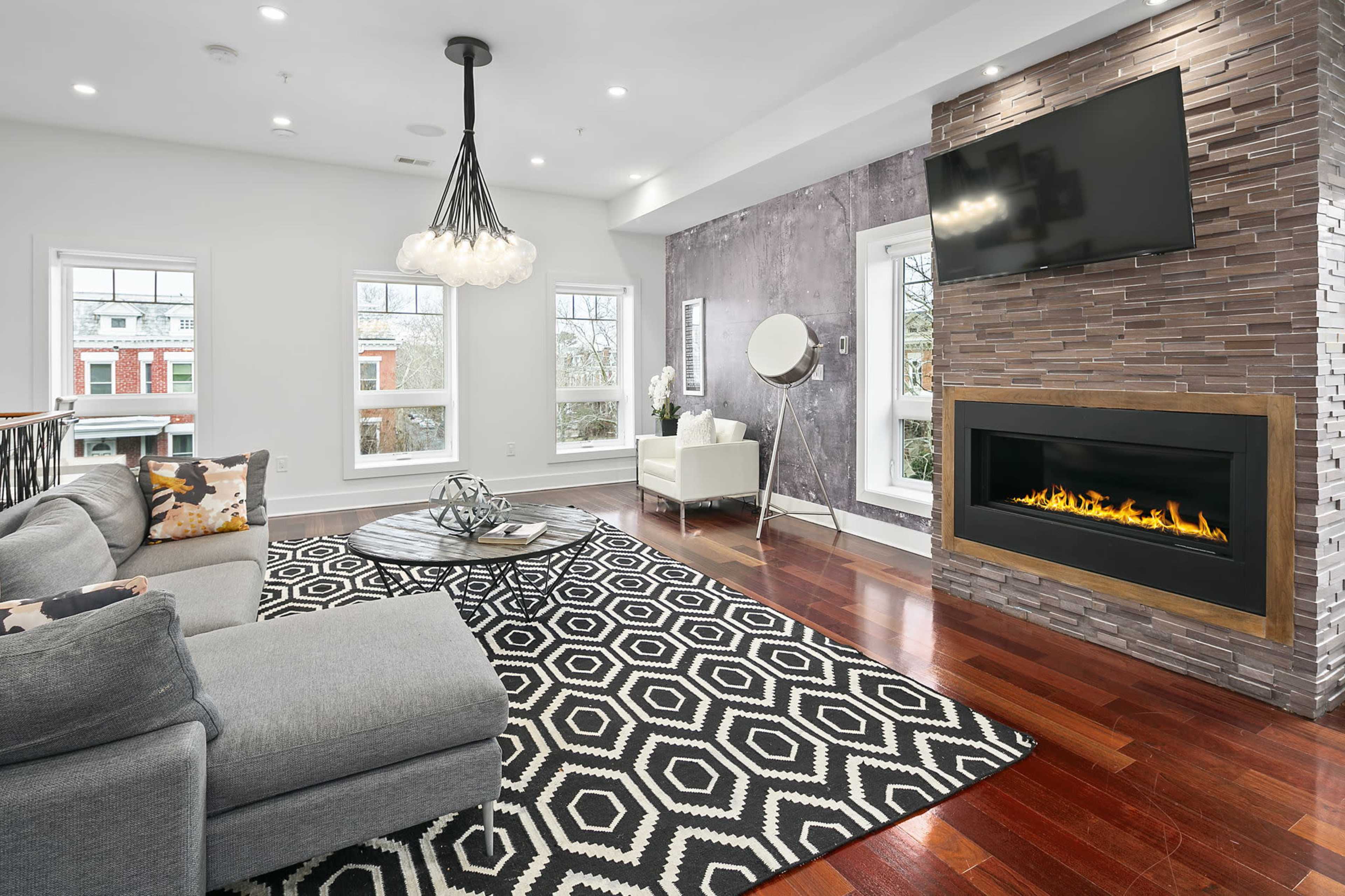 A modern living room features a gray sectional sofa, a geometric black and white rug, a sleek fireplace, and large windows allowing natural light.