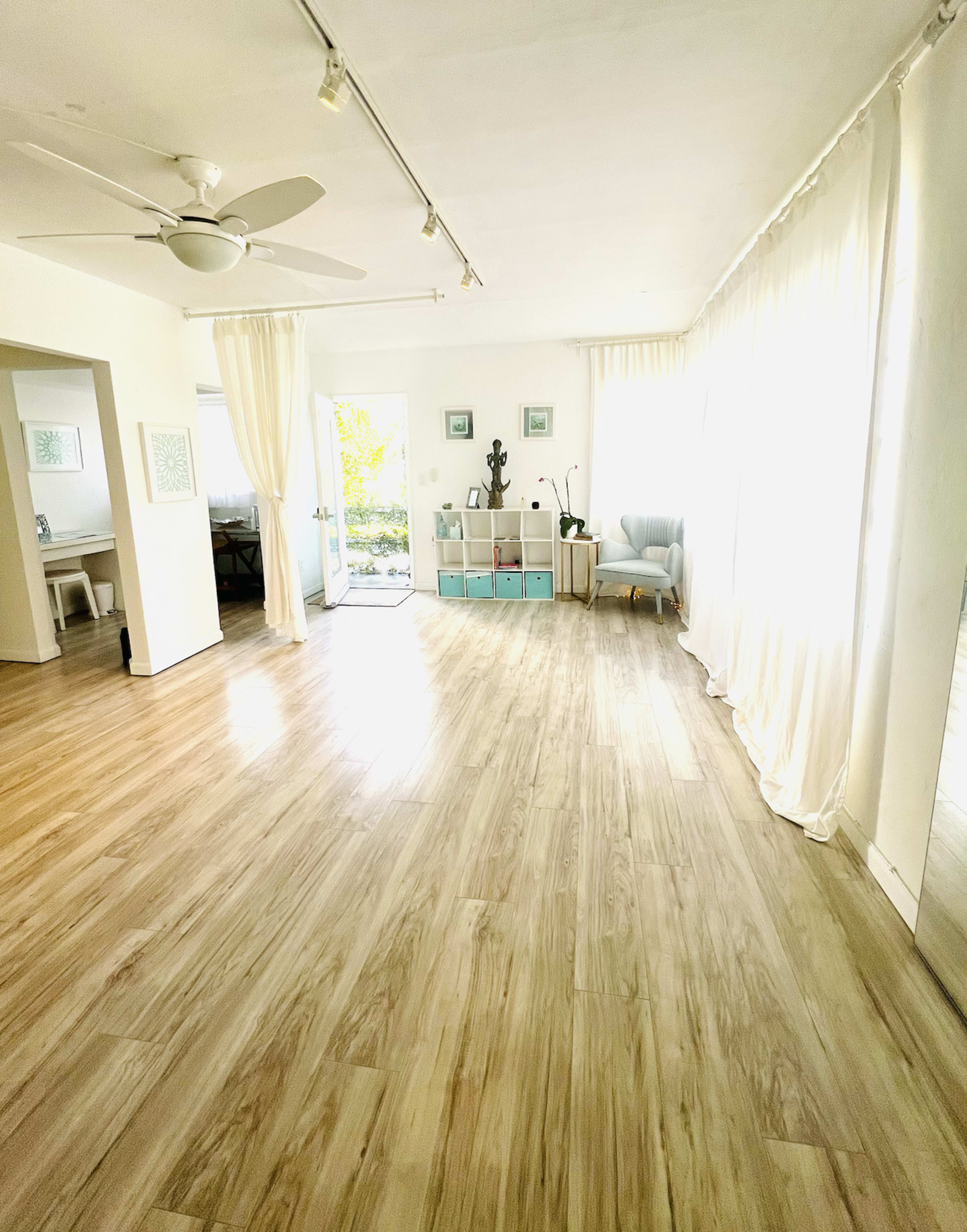 A spacious room with wooden floors, large windows, and a seating area decorated with light curtains and a small bookshelf.