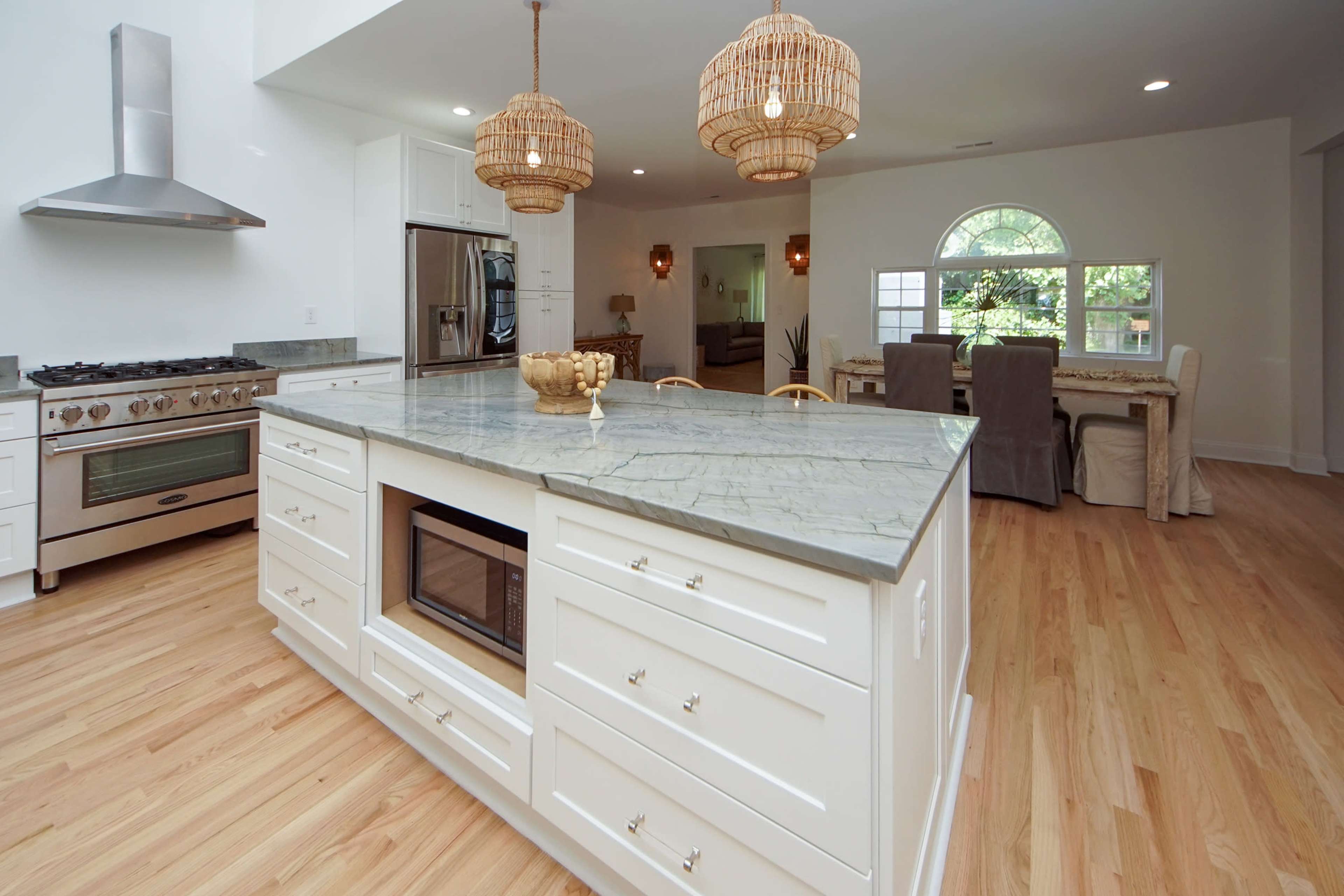 A modern kitchen featuring a large central island with a green marble countertop, white cabinetry, and three woven pendant lights hanging above.