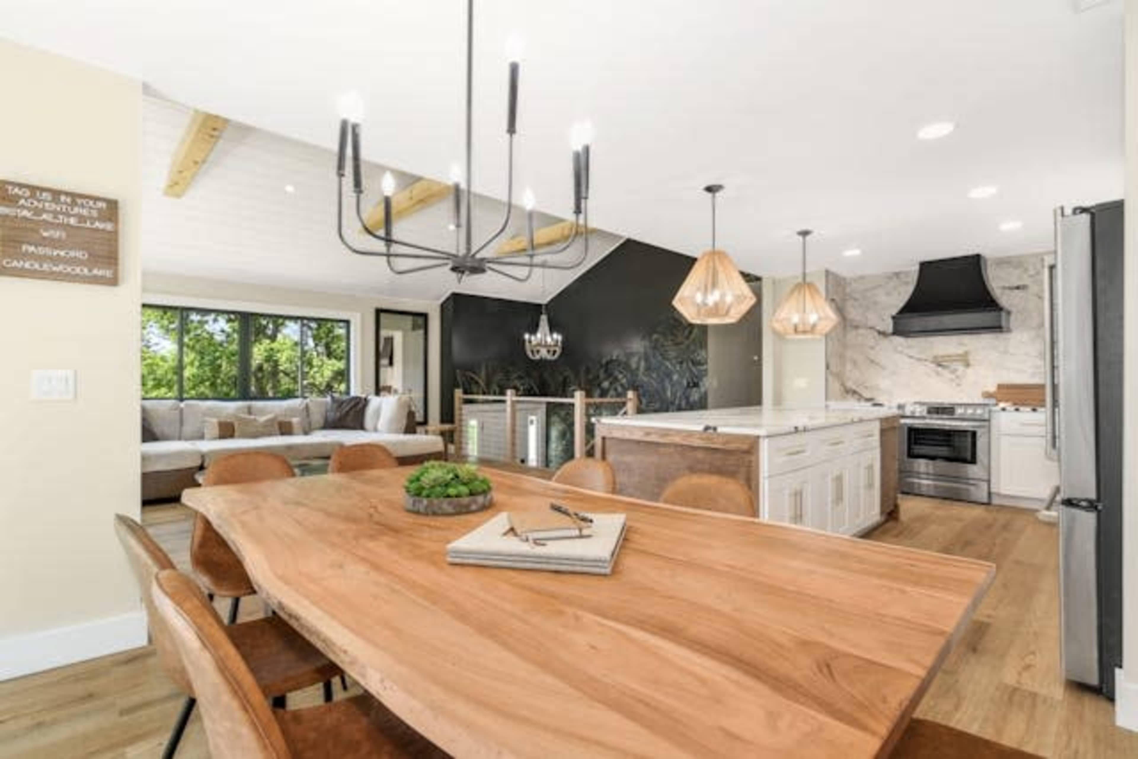 The image shows a modern kitchen and dining area featuring a large wooden table, pendant lights, and a kitchen island with a marble backsplash.