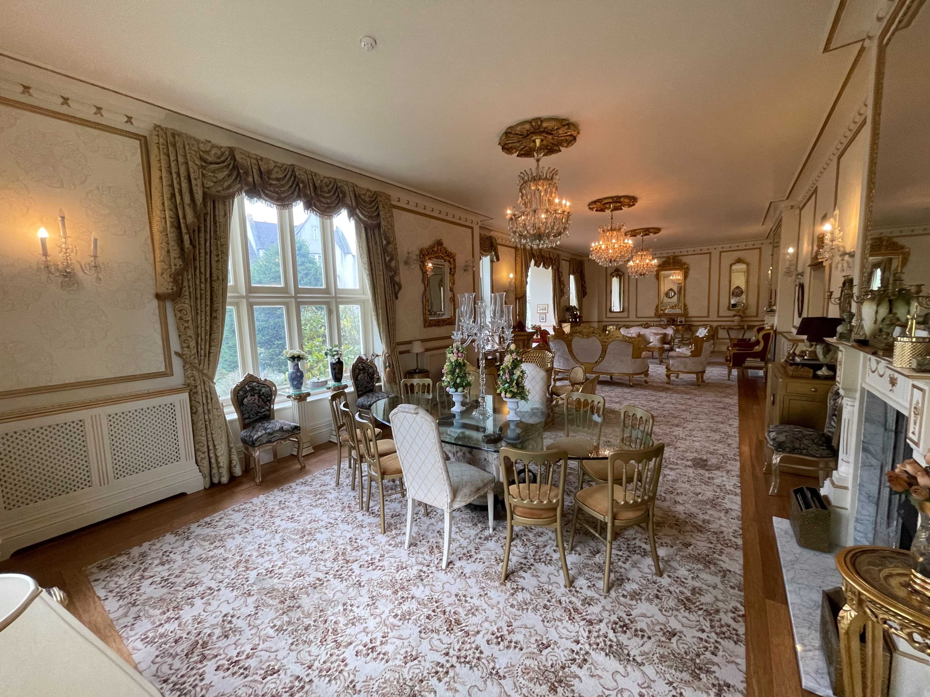 The image shows an elegantly decorated dining room featuring a large round table surrounded by chairs, ornate chandeliers, and plush seating arrangements against a backdrop of elaborate wallpaper and curtains.