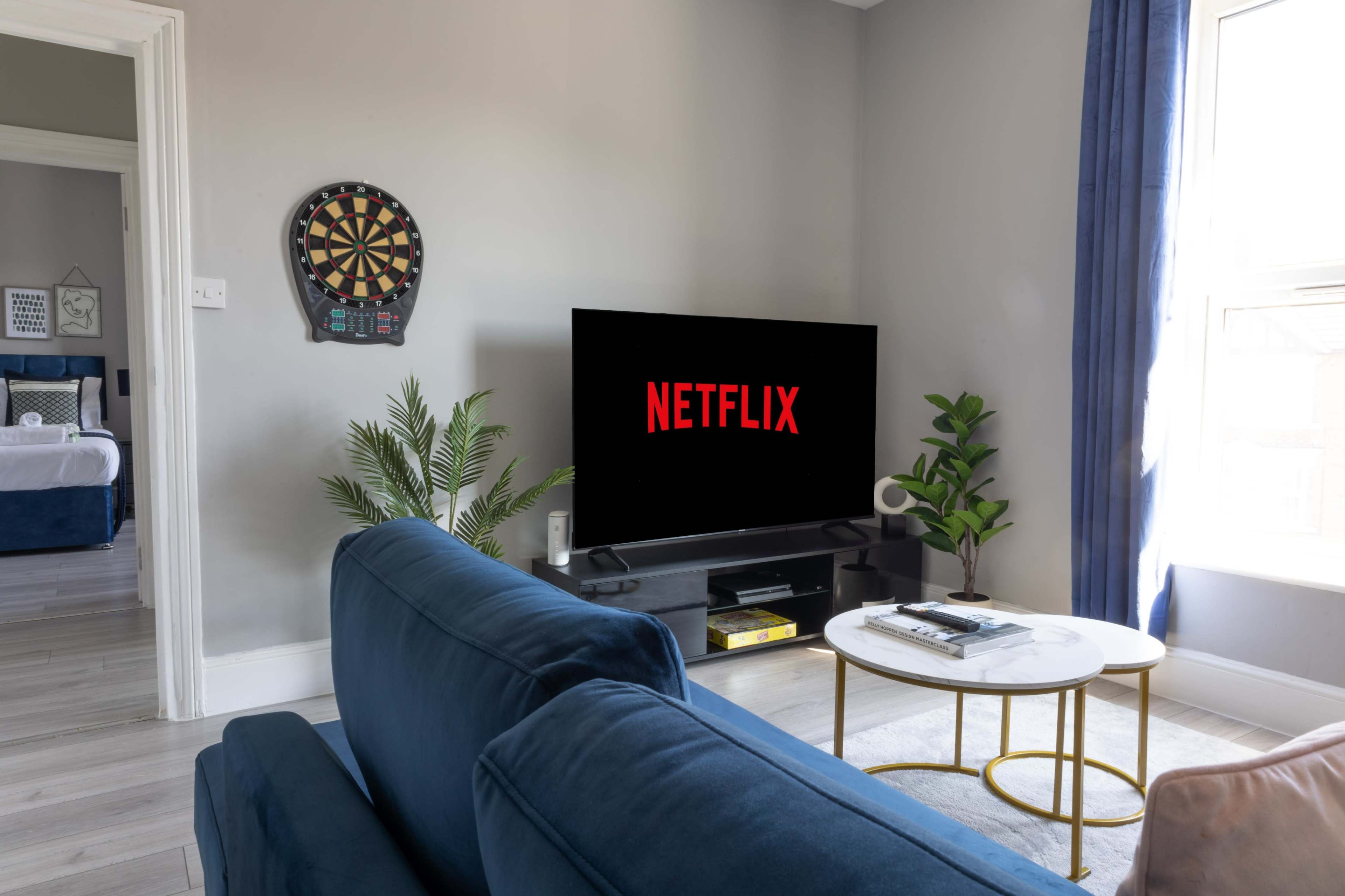 A living room features a large television displaying the Netflix logo, with a dartboard on the wall and a coffee table in front of a blue sofa.