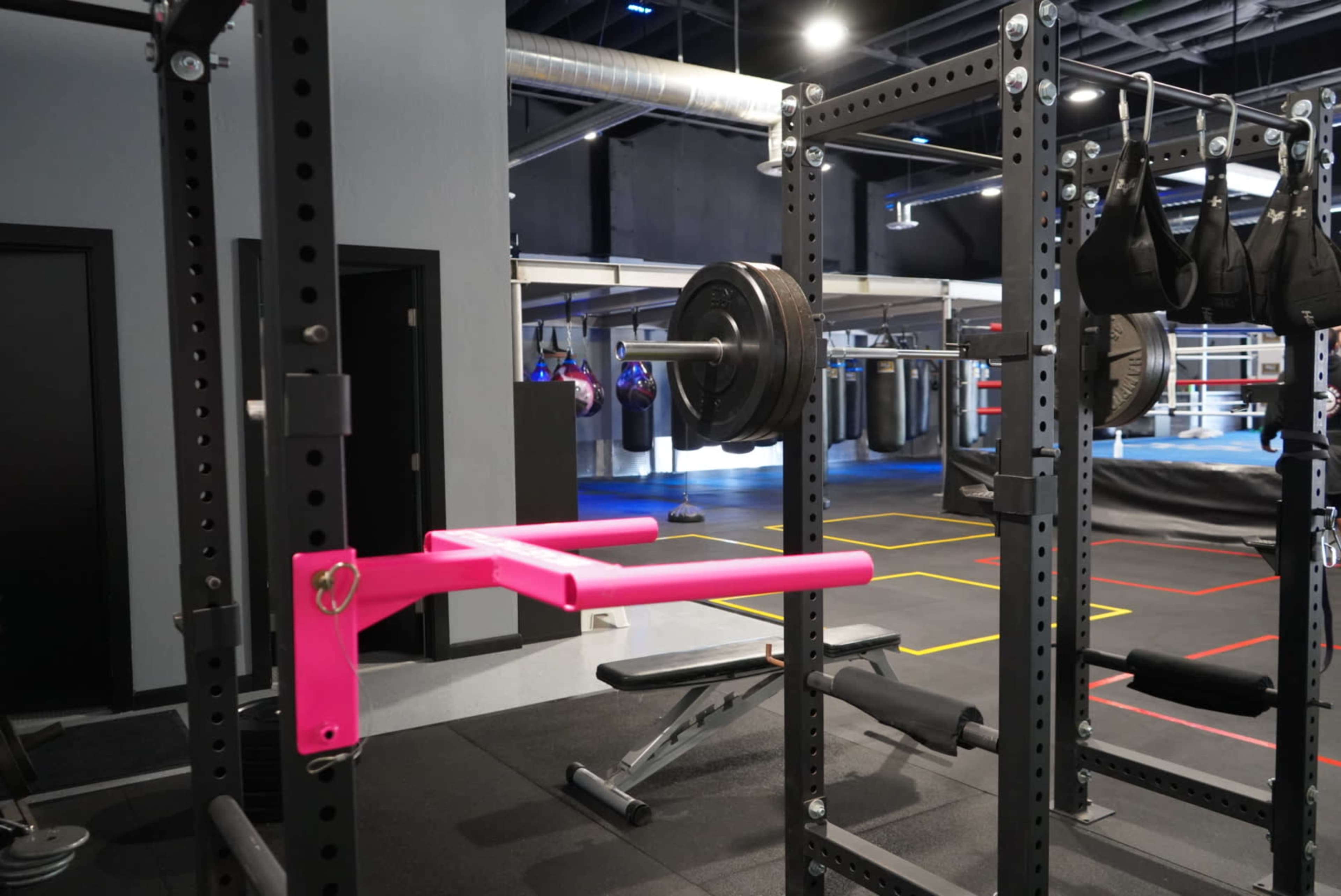 A gym interior with weightlifting equipment, including a barbell on a squat rack and a pink pull-up bar.
