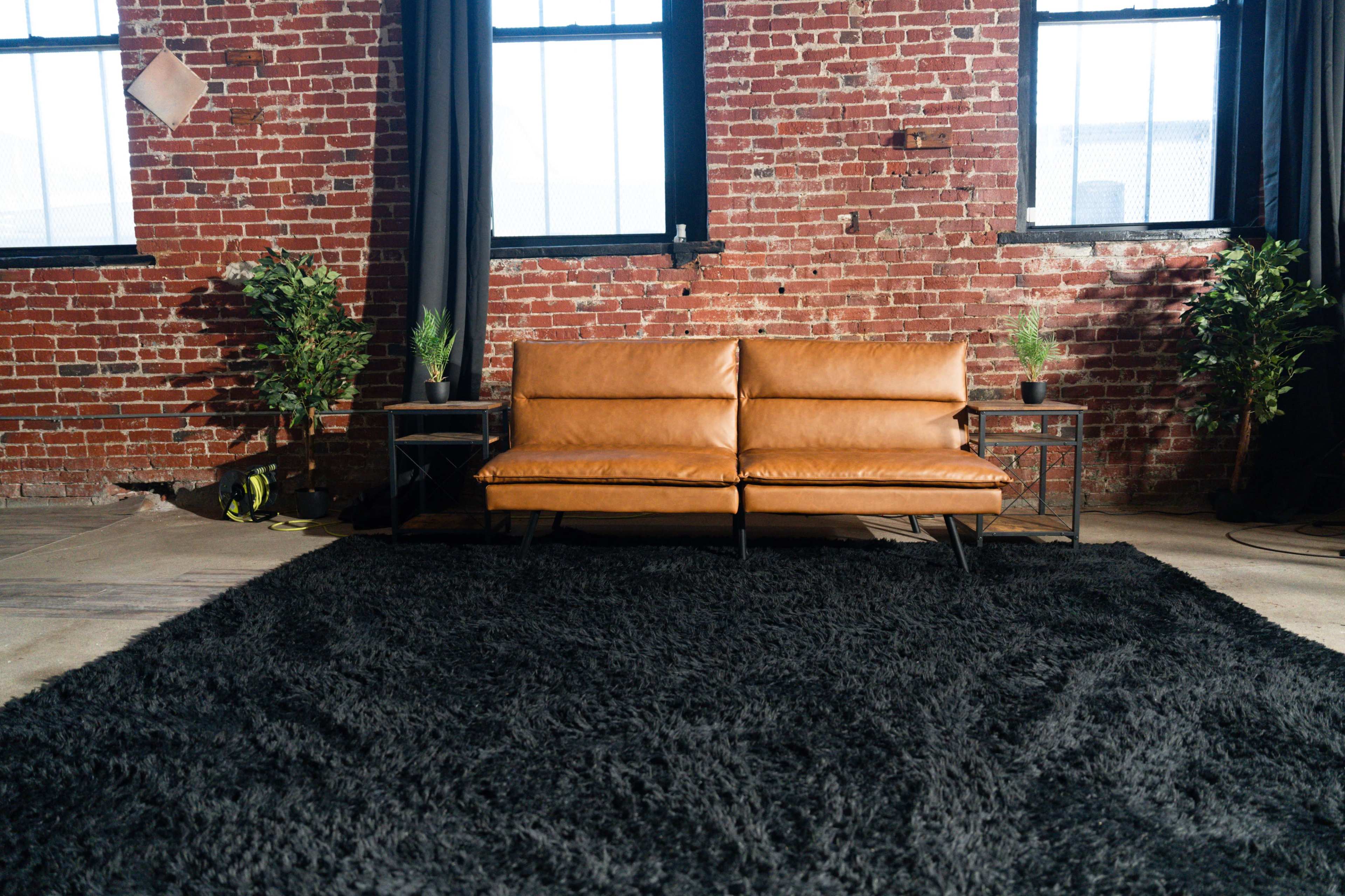 A brown leather couch is positioned in front of a brick wall, surrounded by two small tables and potted plants on either side, with a large black rug covering the floor.