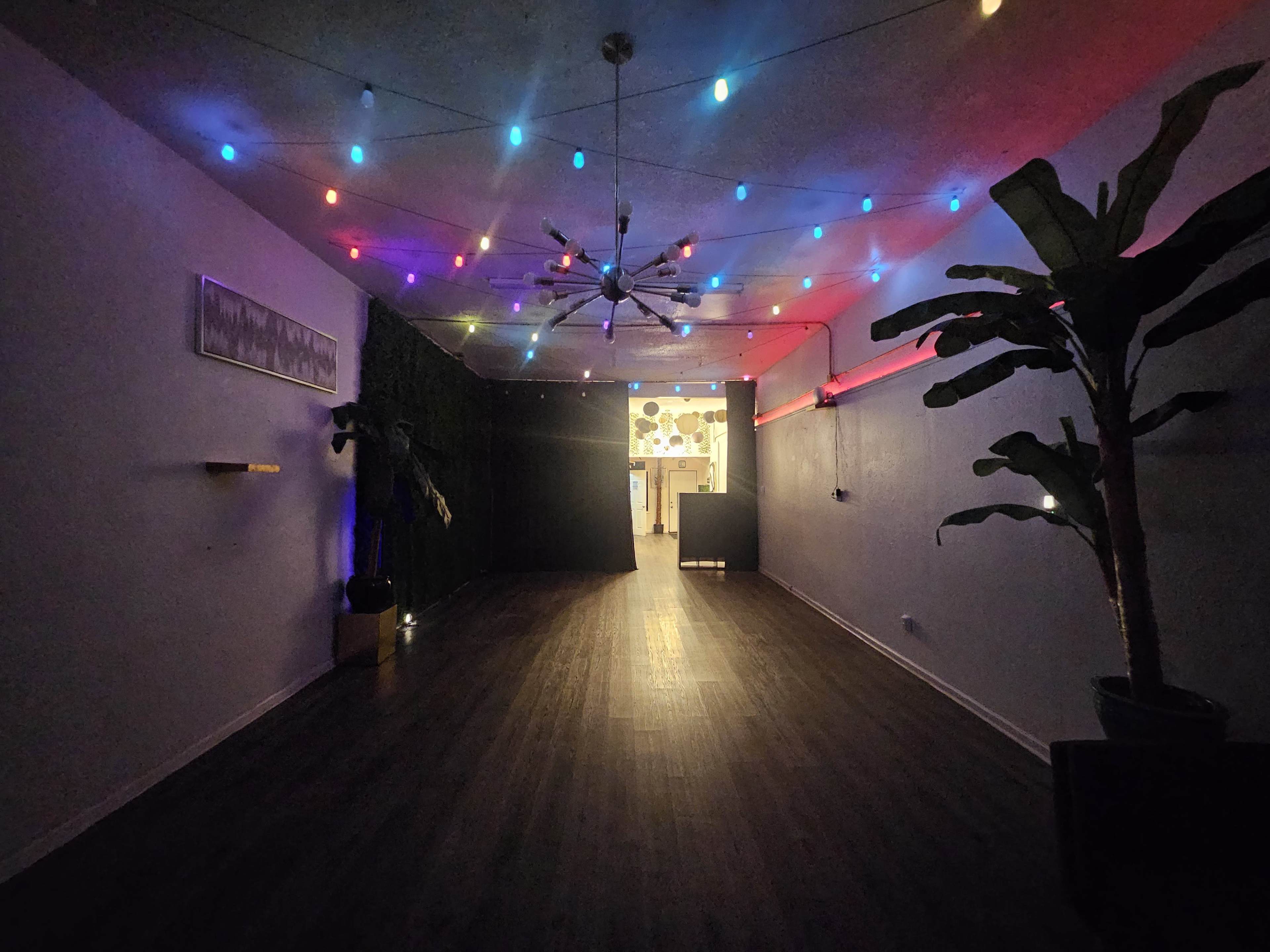 A brightly lit indoor space features colorful string lights and a plant, leading to a darkened entryway at the far end.