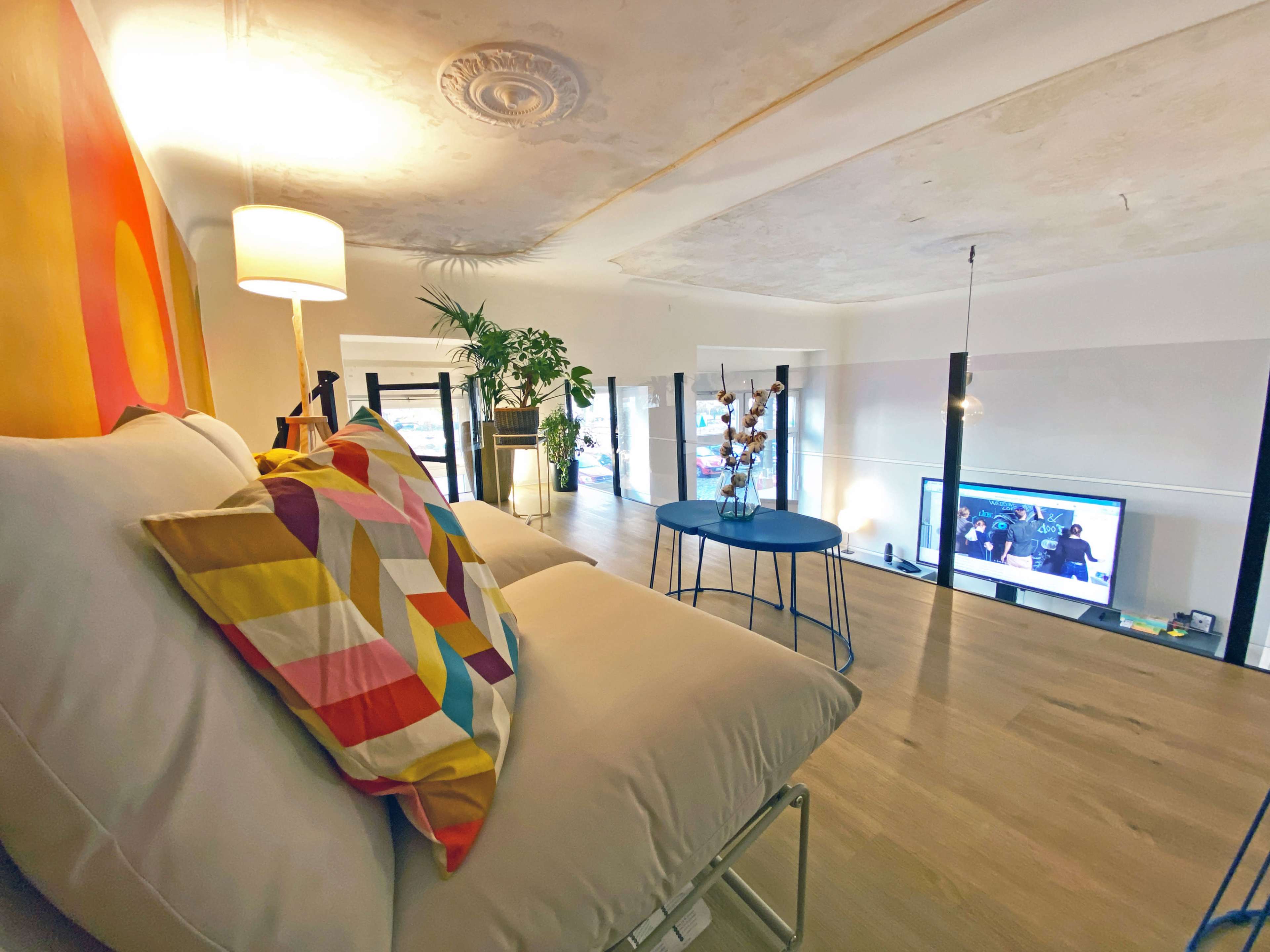 A modern, airy loft with a colorful pillow arrangement on a bed and a television visible in the background.