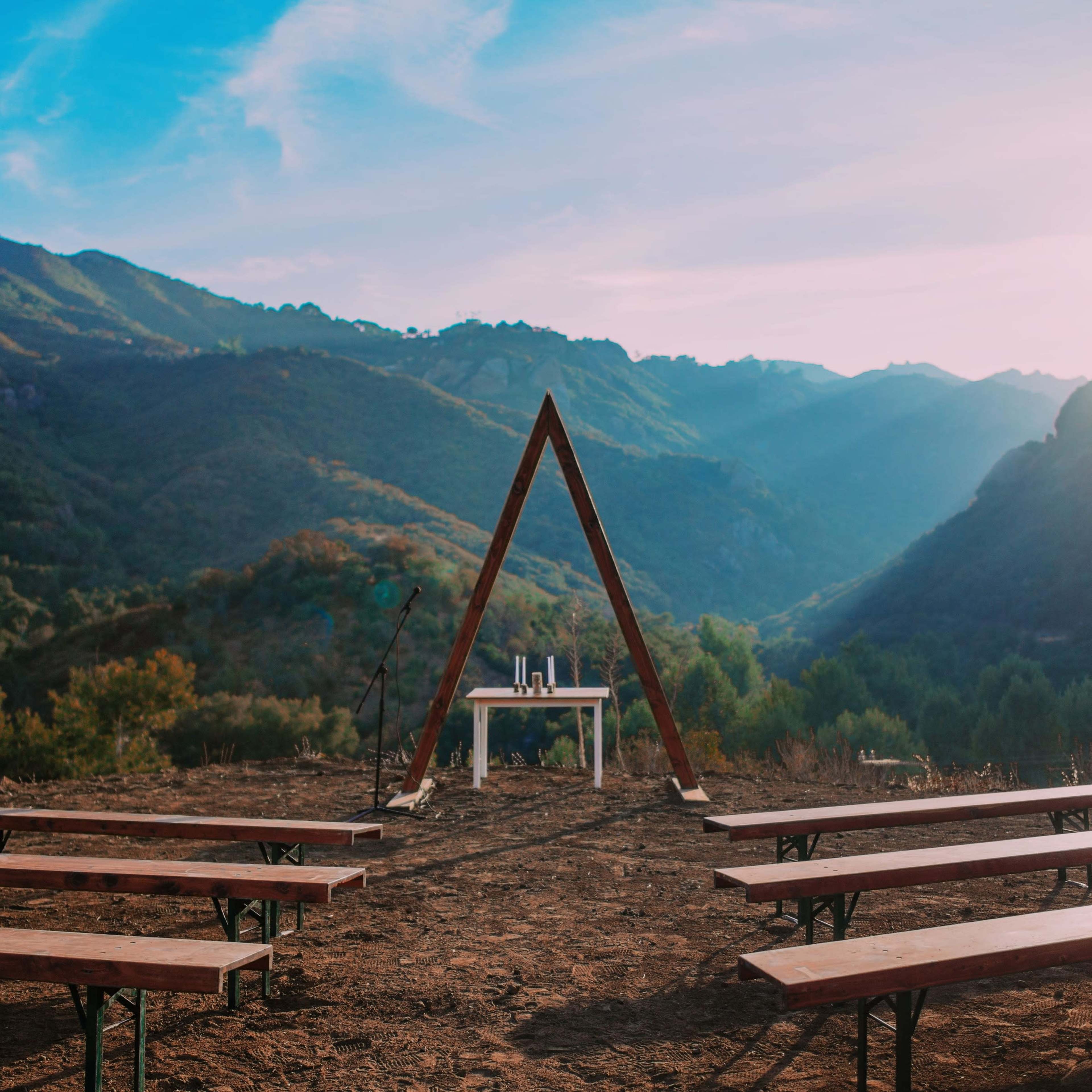A wooden triangular arch stands behind a table in an outdoor setting surrounded by mountains and rows of benches.