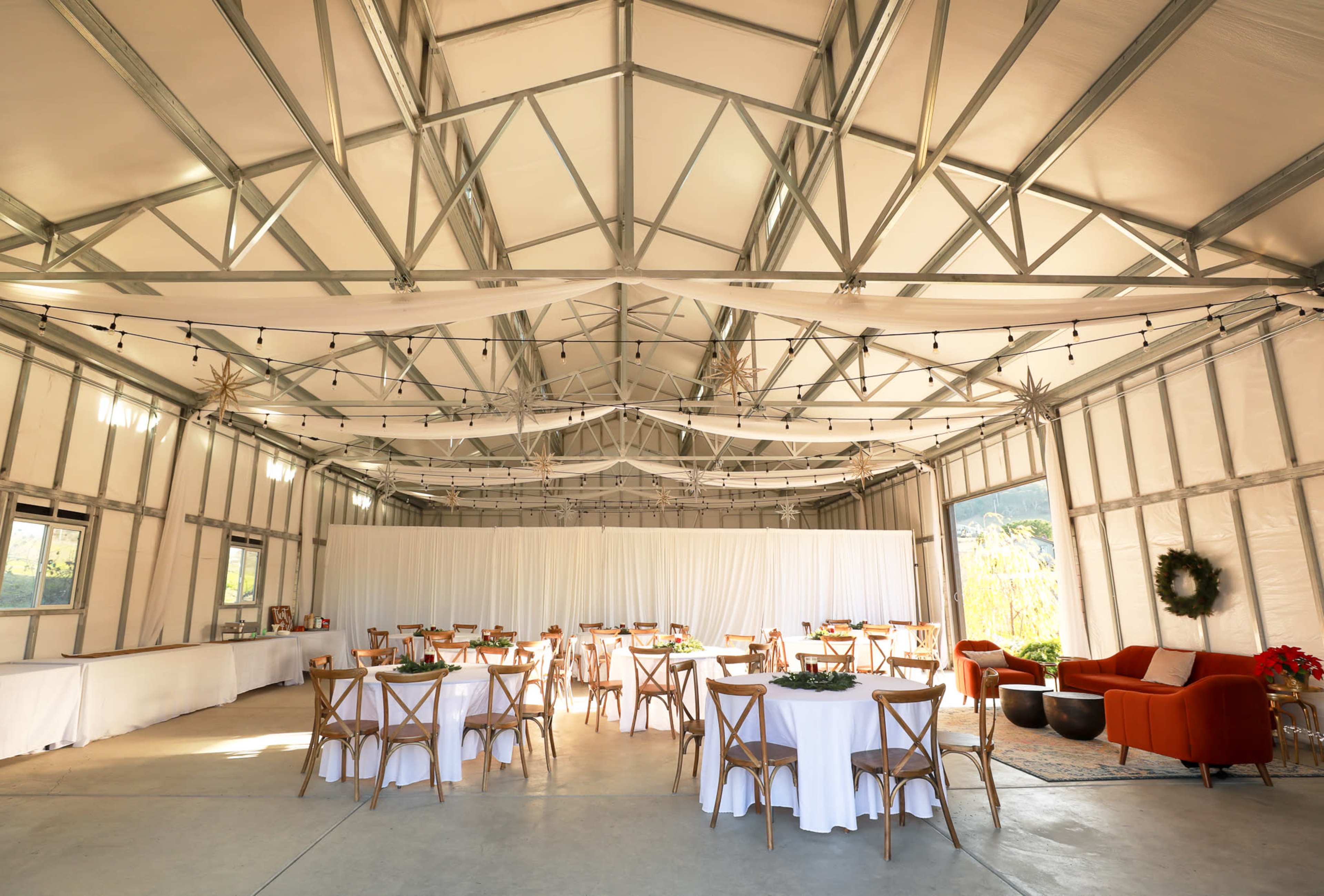 A spacious indoor venue features round tables with white tablecloths, surrounded by wooden chairs, and is illuminated by overhead string lights.