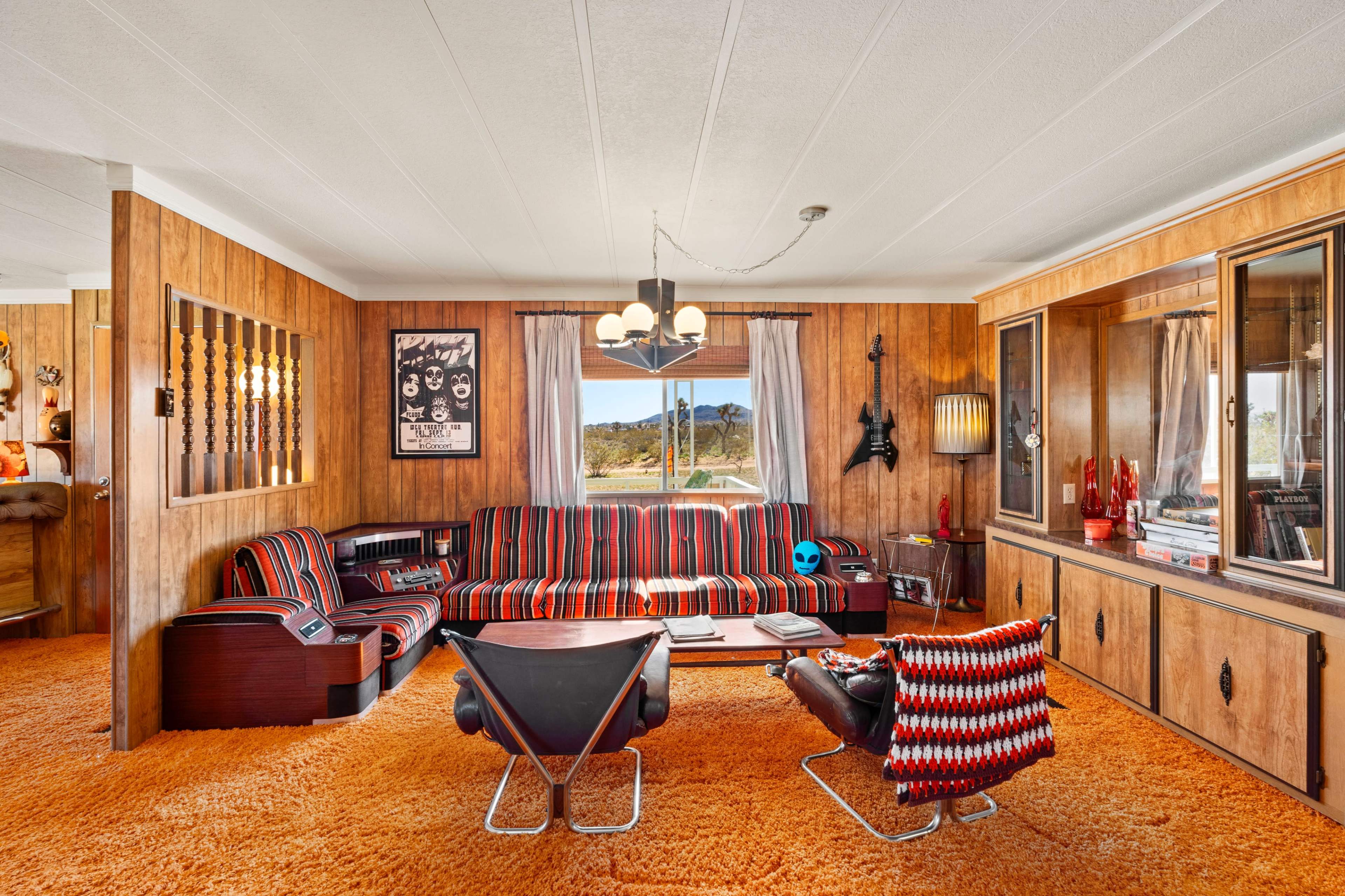 A retro living room with wood-paneled walls, orange carpet, striped furniture, and large windows overlooking a landscape.