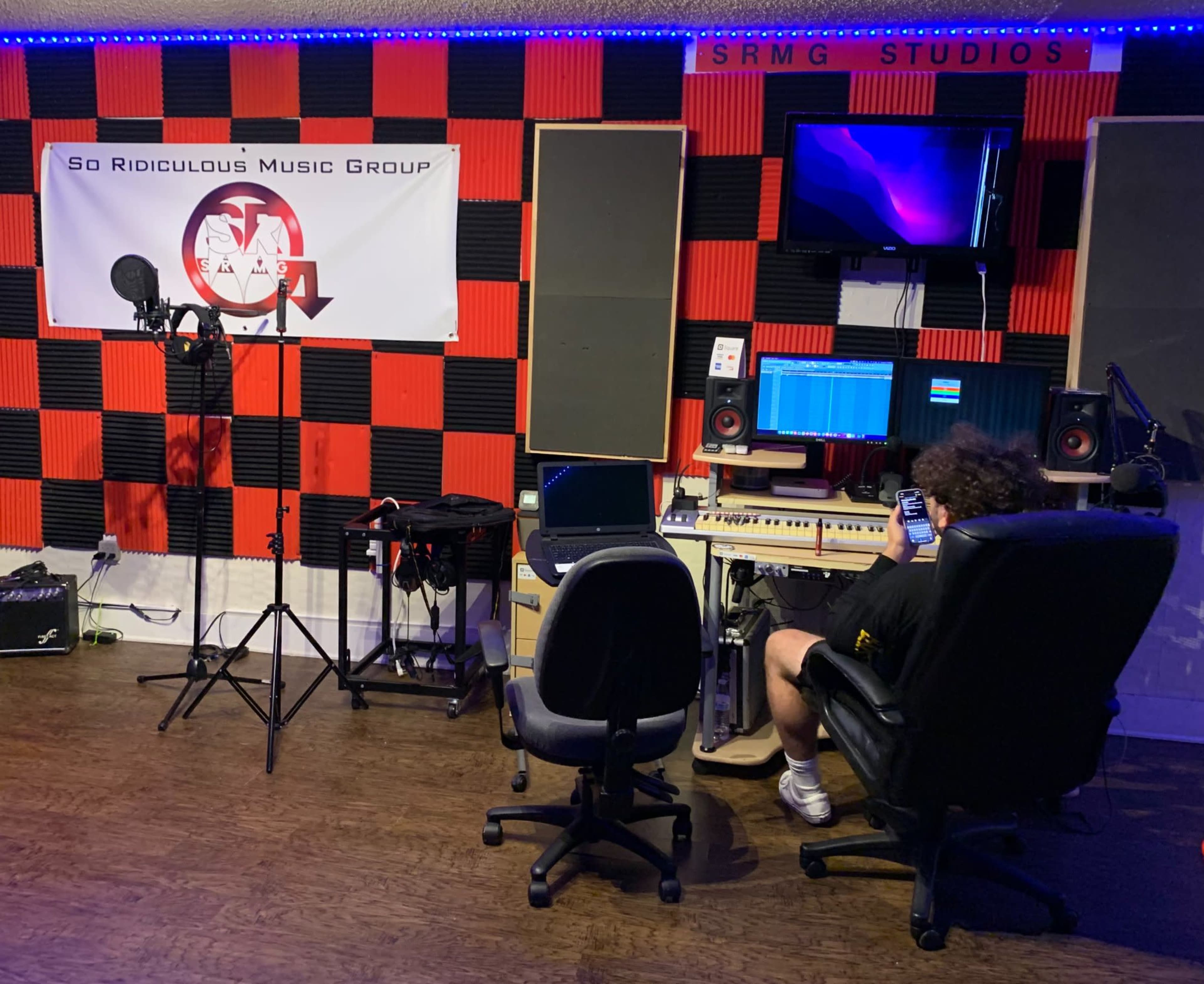 A person sits on a chair using a phone in a music studio with black and red soundproofing, microphones, and audio equipment.