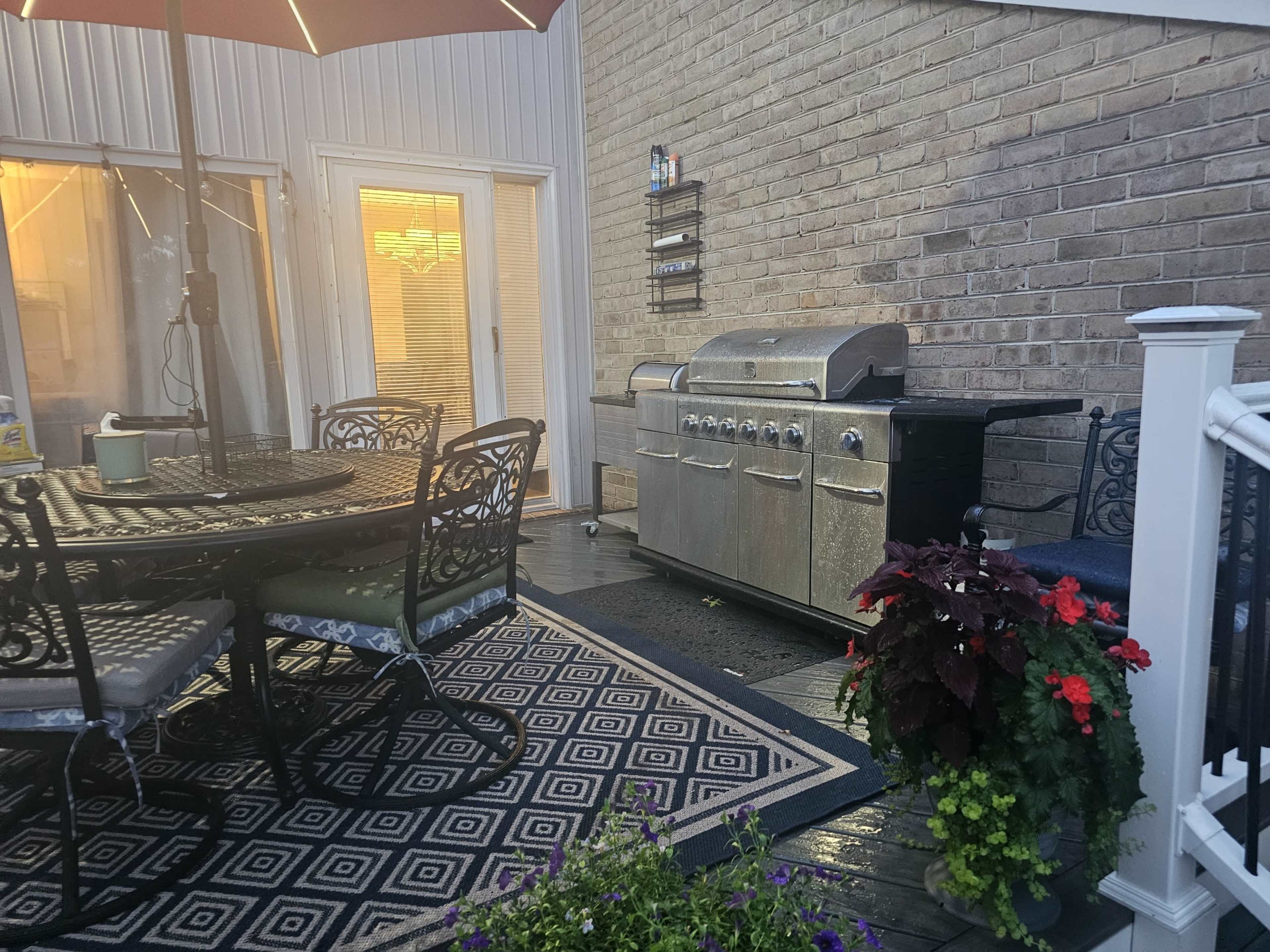 The image shows a backyard patio setup featuring a dining table with chairs, a grill, and potted plants, all situated under an umbrella.