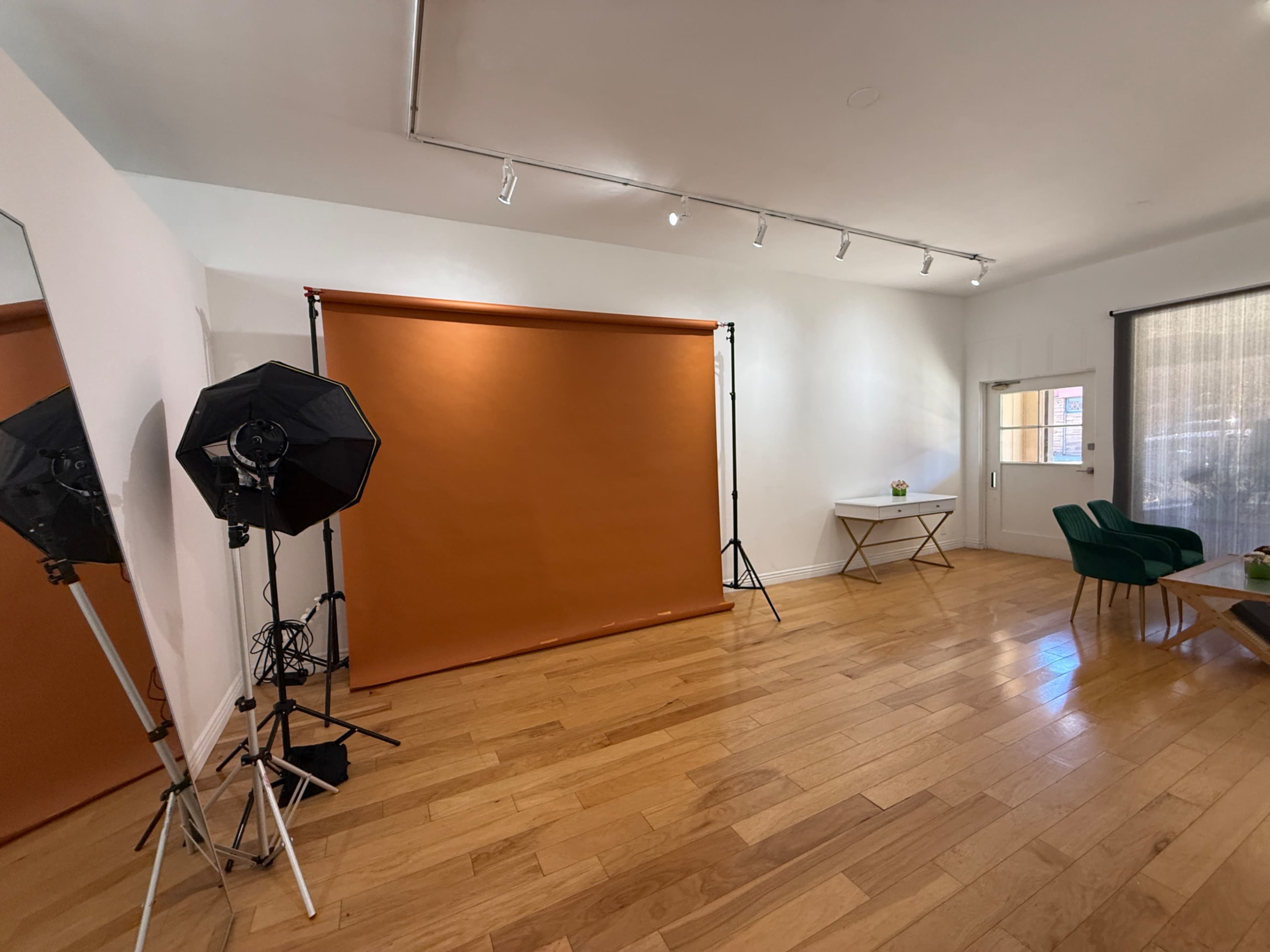 The image shows a well-lit room featuring a brown backdrop set up on stands, two softbox lights, and minimal furnishings including a green chair and a white table.