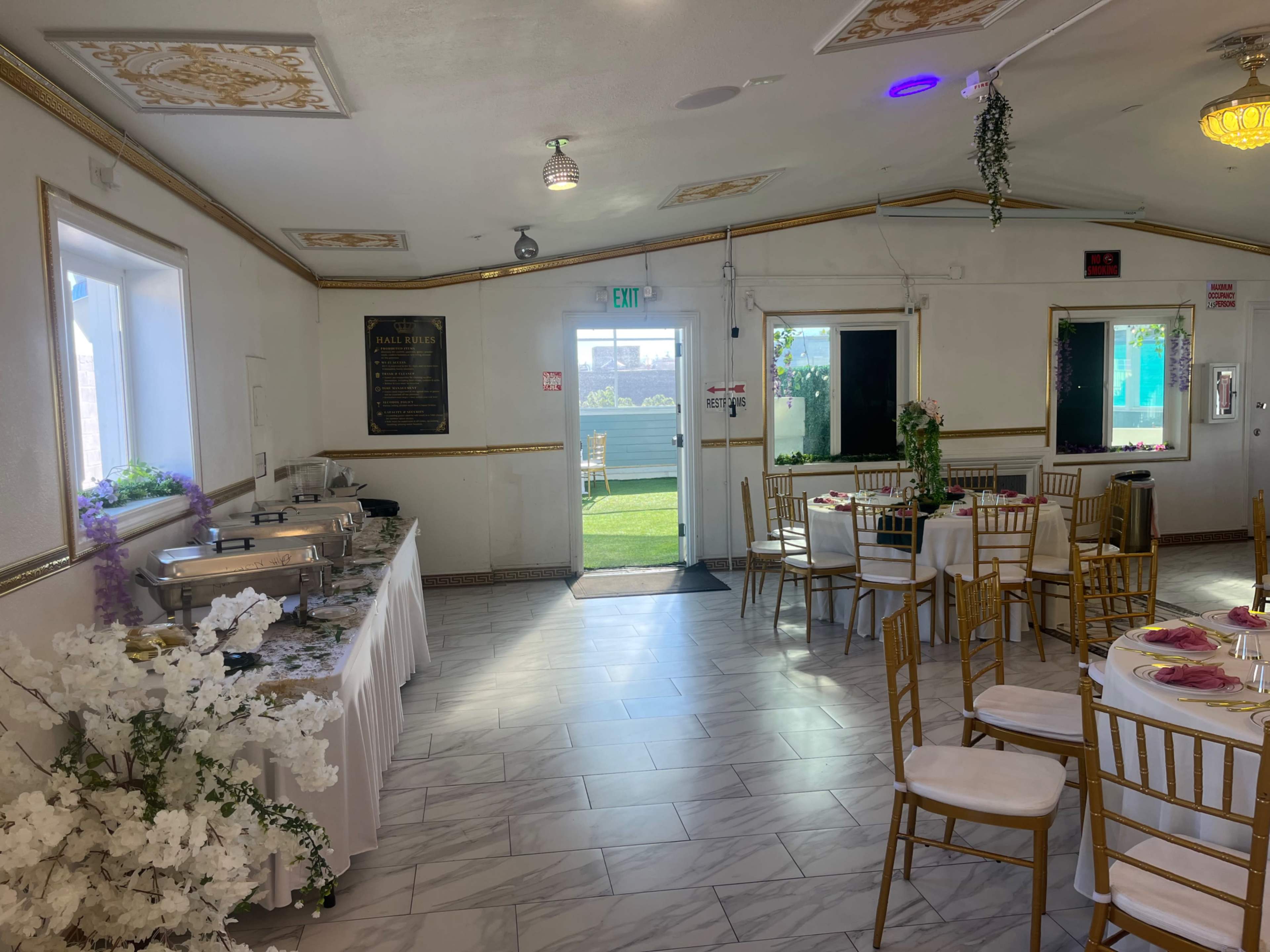 The image shows a decorated room set for dining, featuring tables with white tablecloths, gold chairs, and a buffet table along one wall.