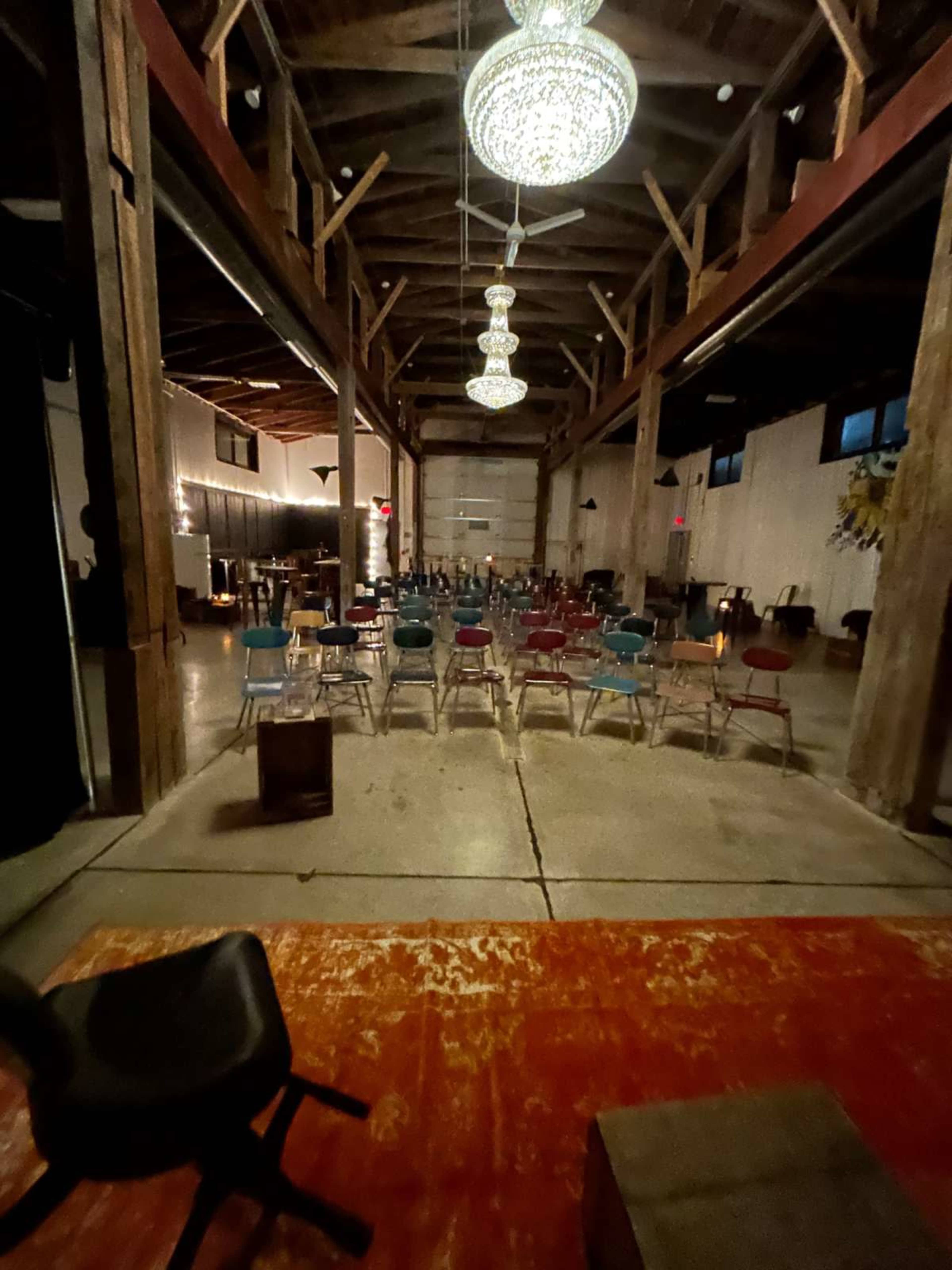 The image shows a spacious, dimly lit venue with rows of colorful chairs arranged for an event, under decorative chandeliers hanging from a wooden ceiling.