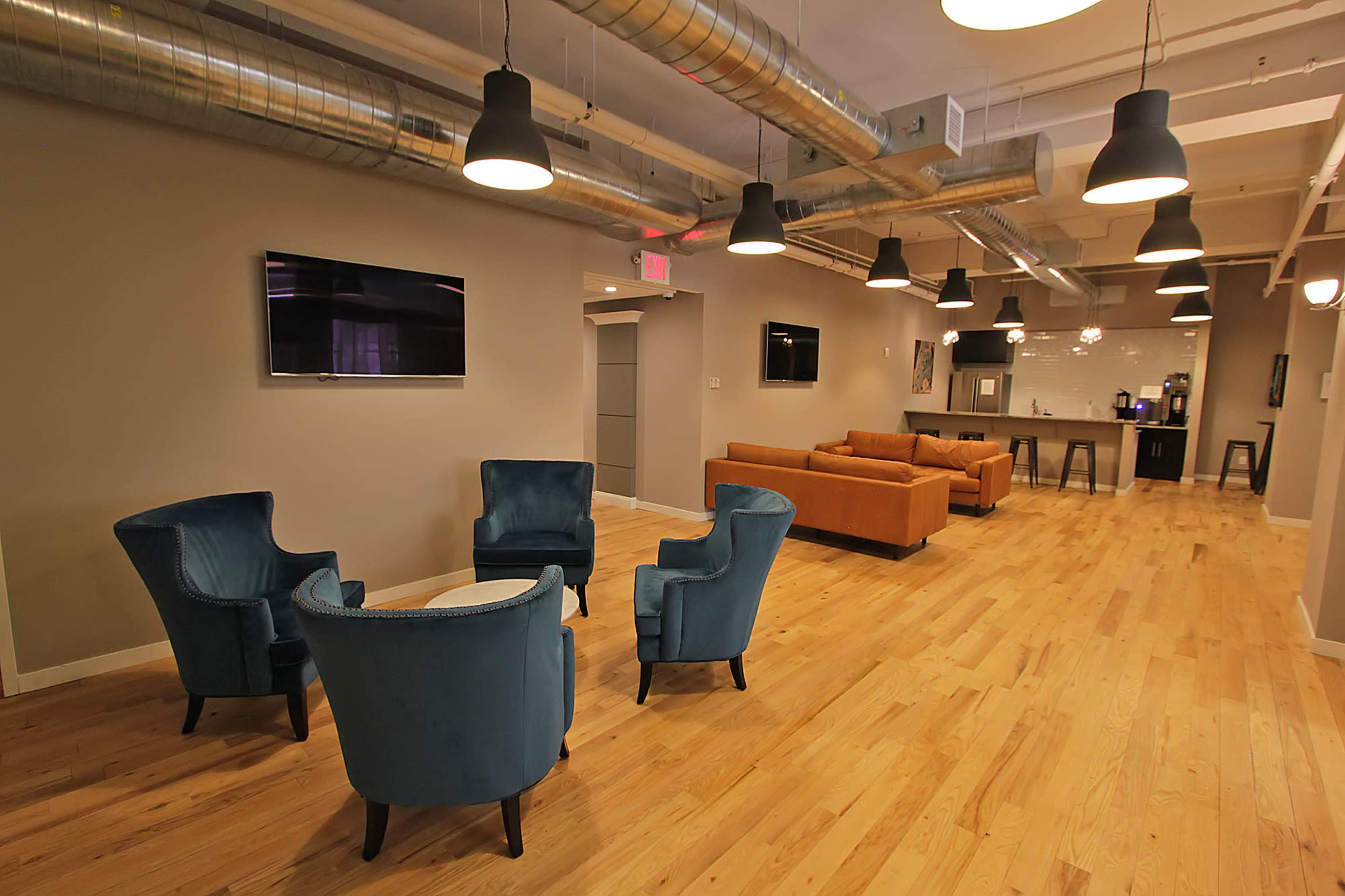 A modern lounge area with two sets of seating, including four blue chairs around a small table and an orange couch, alongside two wall-mounted TVs and a kitchenette in the background.