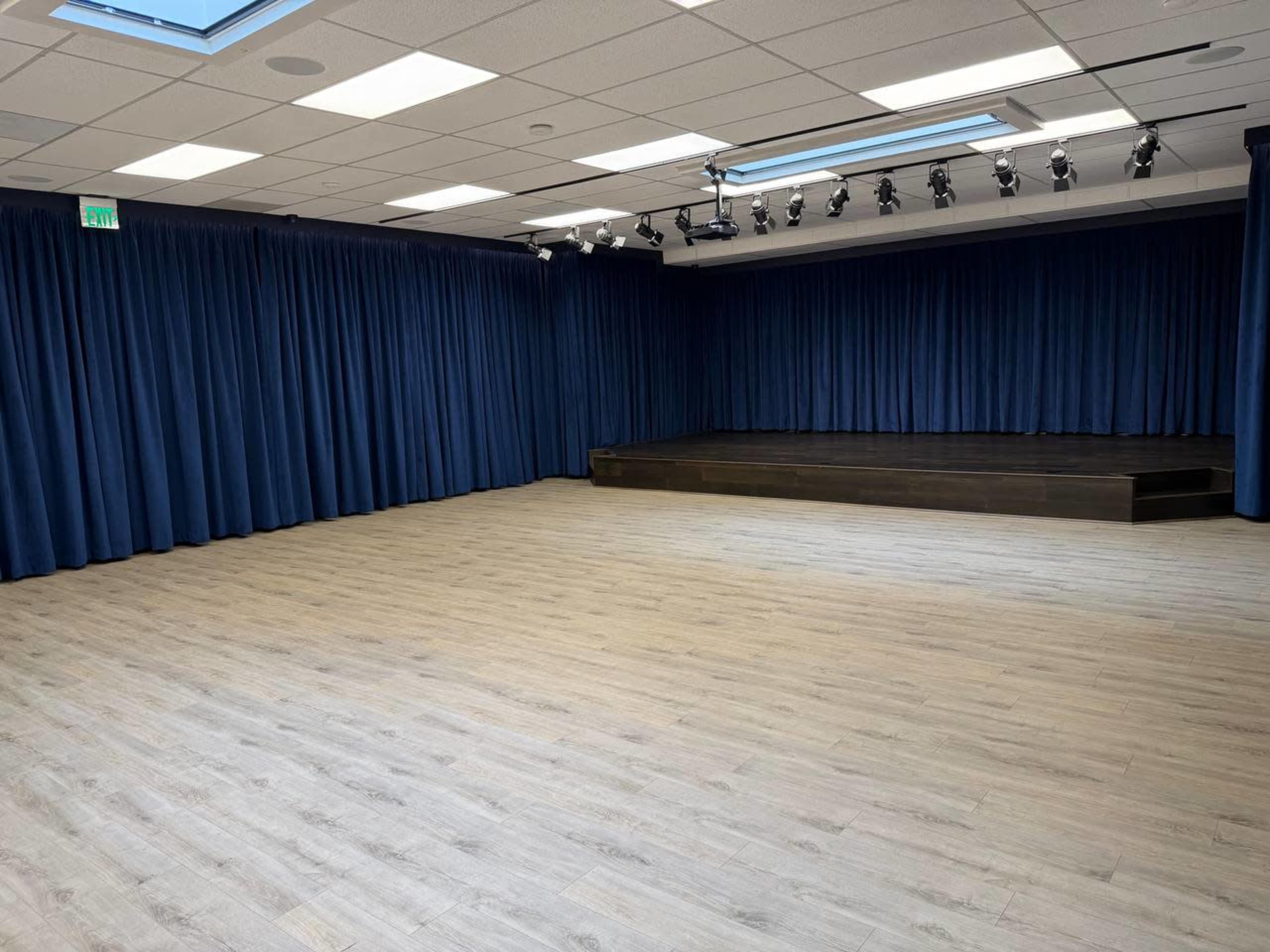 The image shows a spacious, empty room with wooden flooring, blue curtains along the walls, and a small elevated stage at the back.