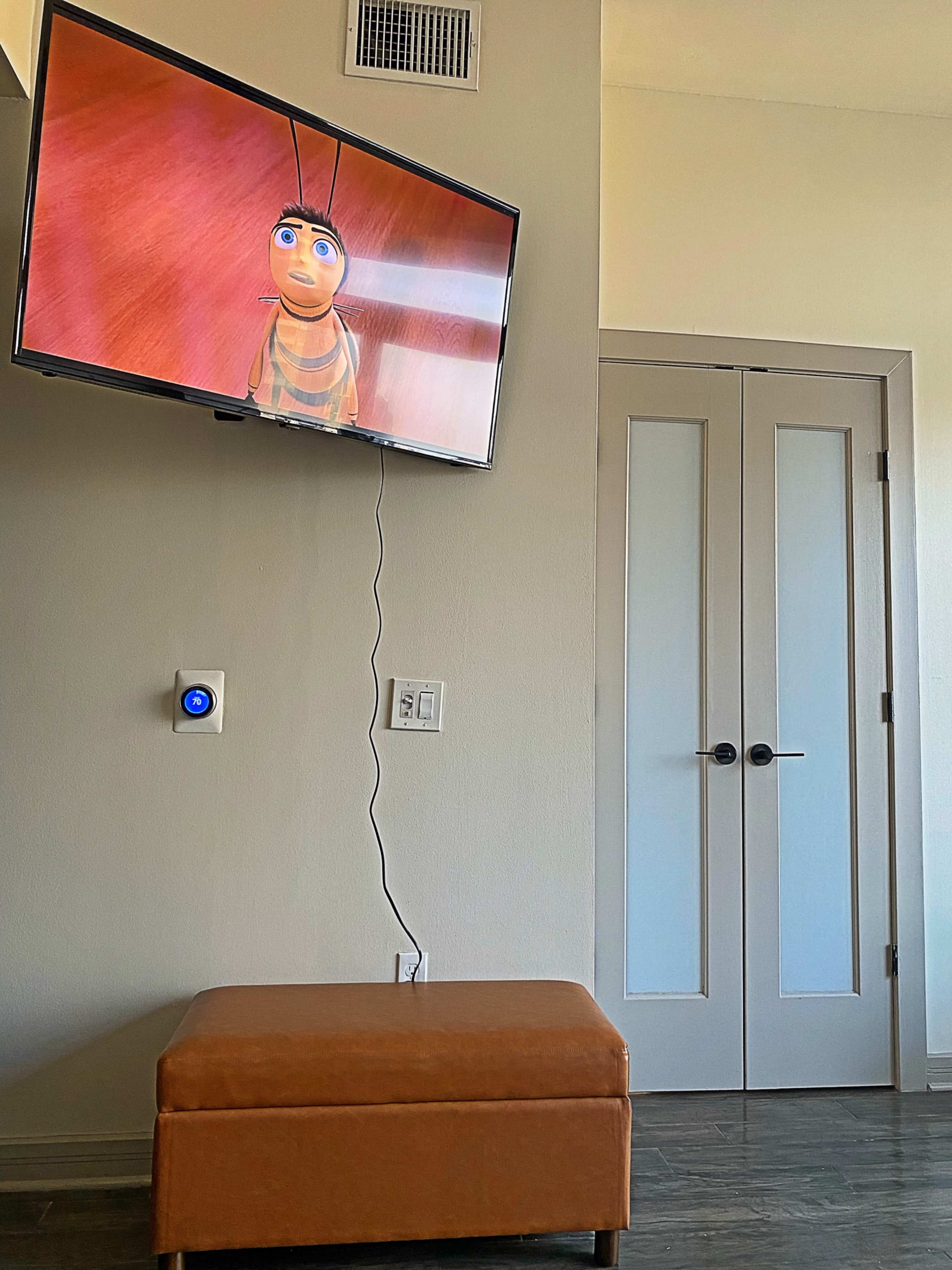 A television displaying an animated character is mounted on a wall above a brown ottoman, beside a set of double doors.