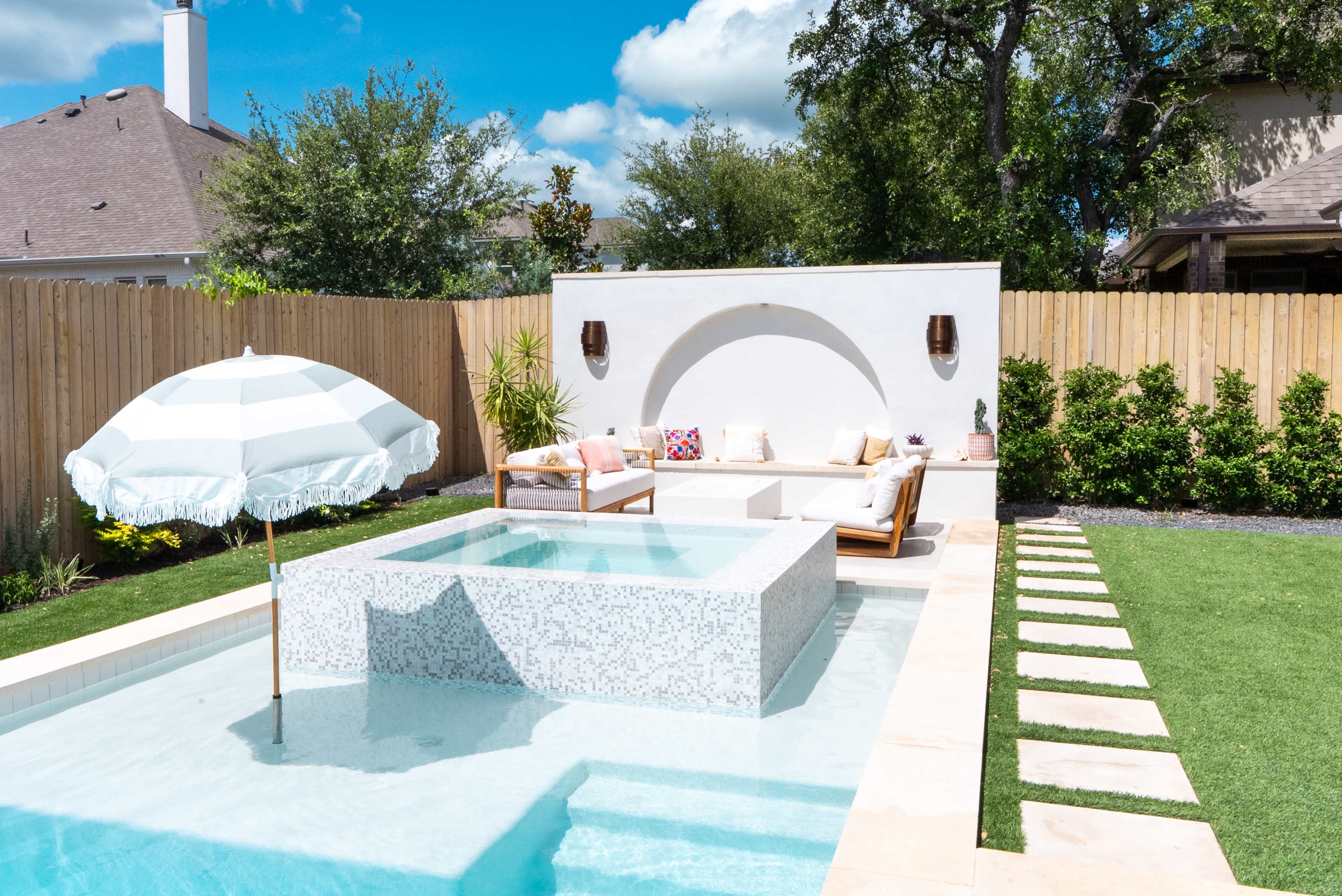 The image shows a modern backyard featuring a swimming pool with a mosaic centerpiece, a shaded seating area, and landscaped grass and plants.
