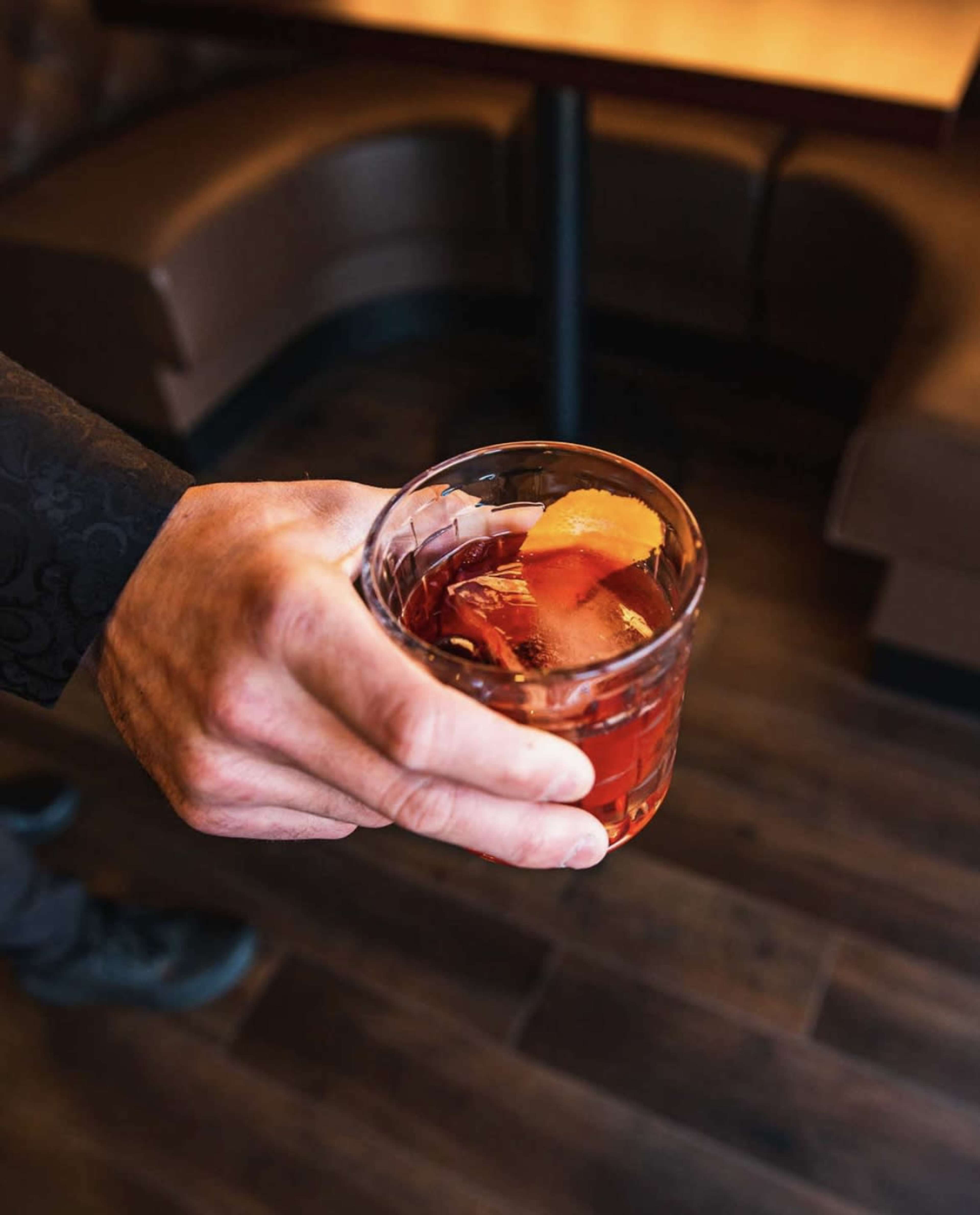 A hand holds a glass of a cocktail with ice and an orange peel garnish in a dimly lit setting.
