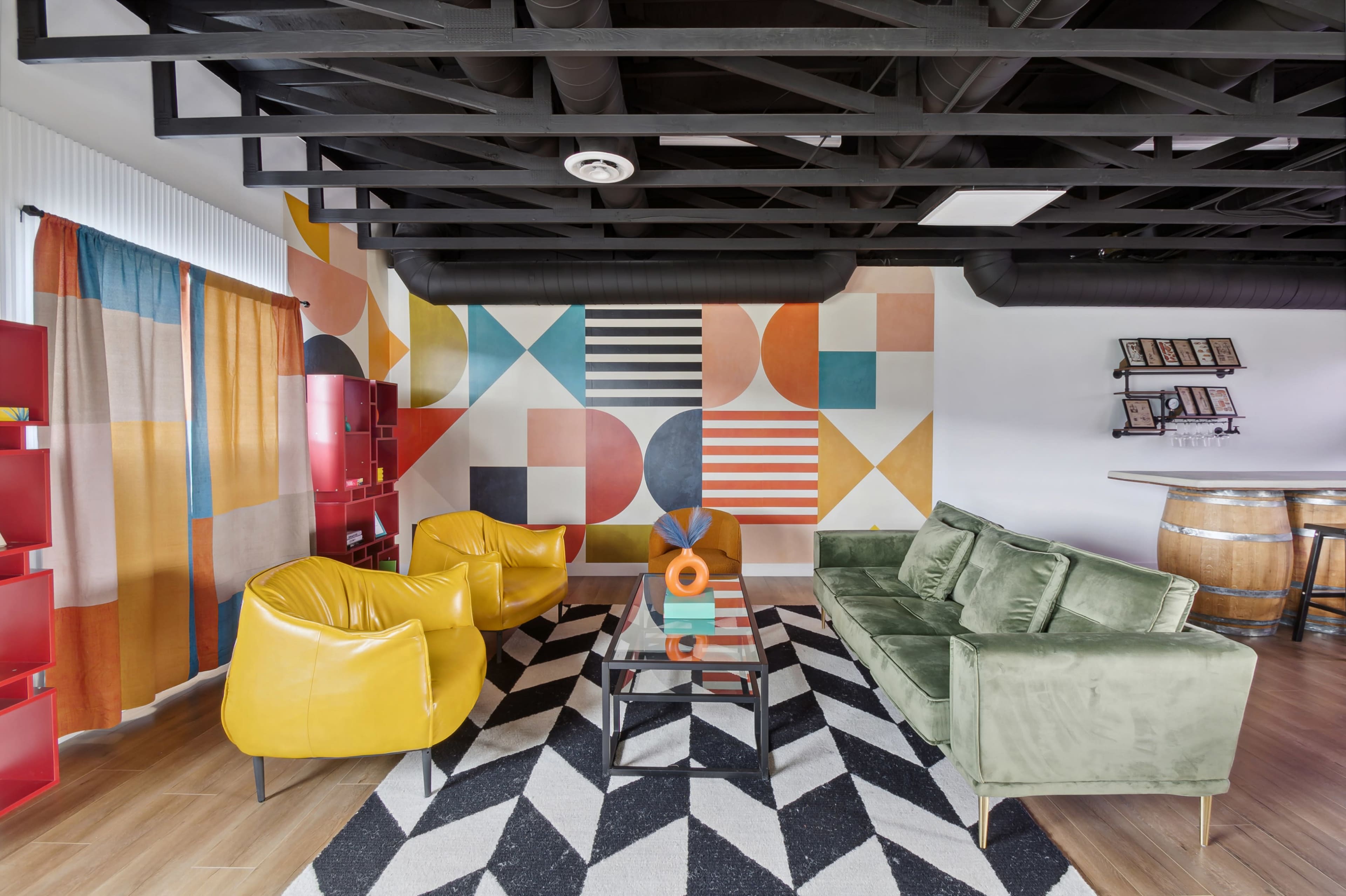 The image shows a modern lounge area with colorful patterned walls, yellow and green seating, a geometric black and white rug, and wooden accents.