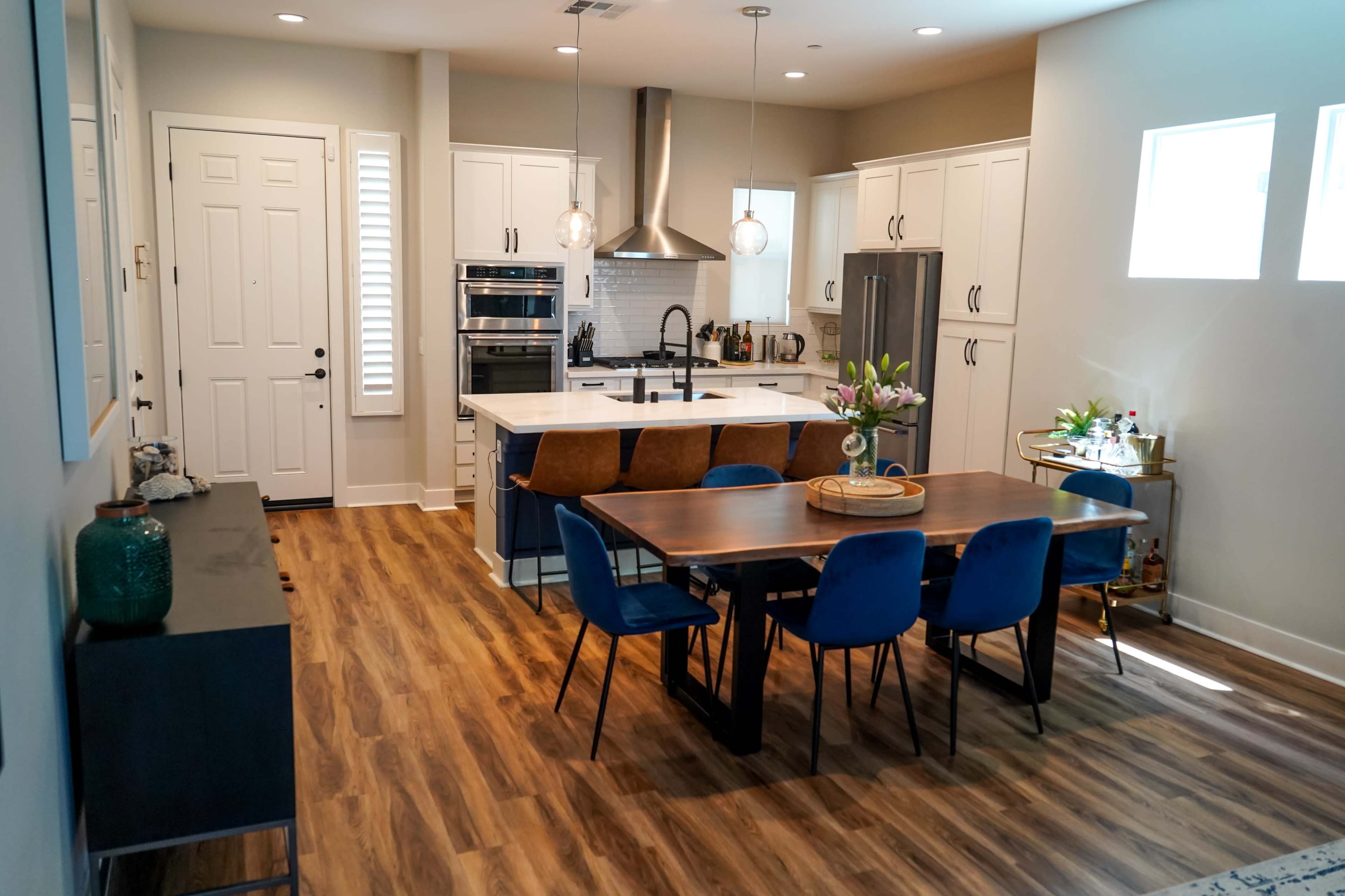 A modern kitchen and dining area featuring white cabinetry, stainless steel appliances, a large island, and a wooden dining table surrounded by blue chairs.