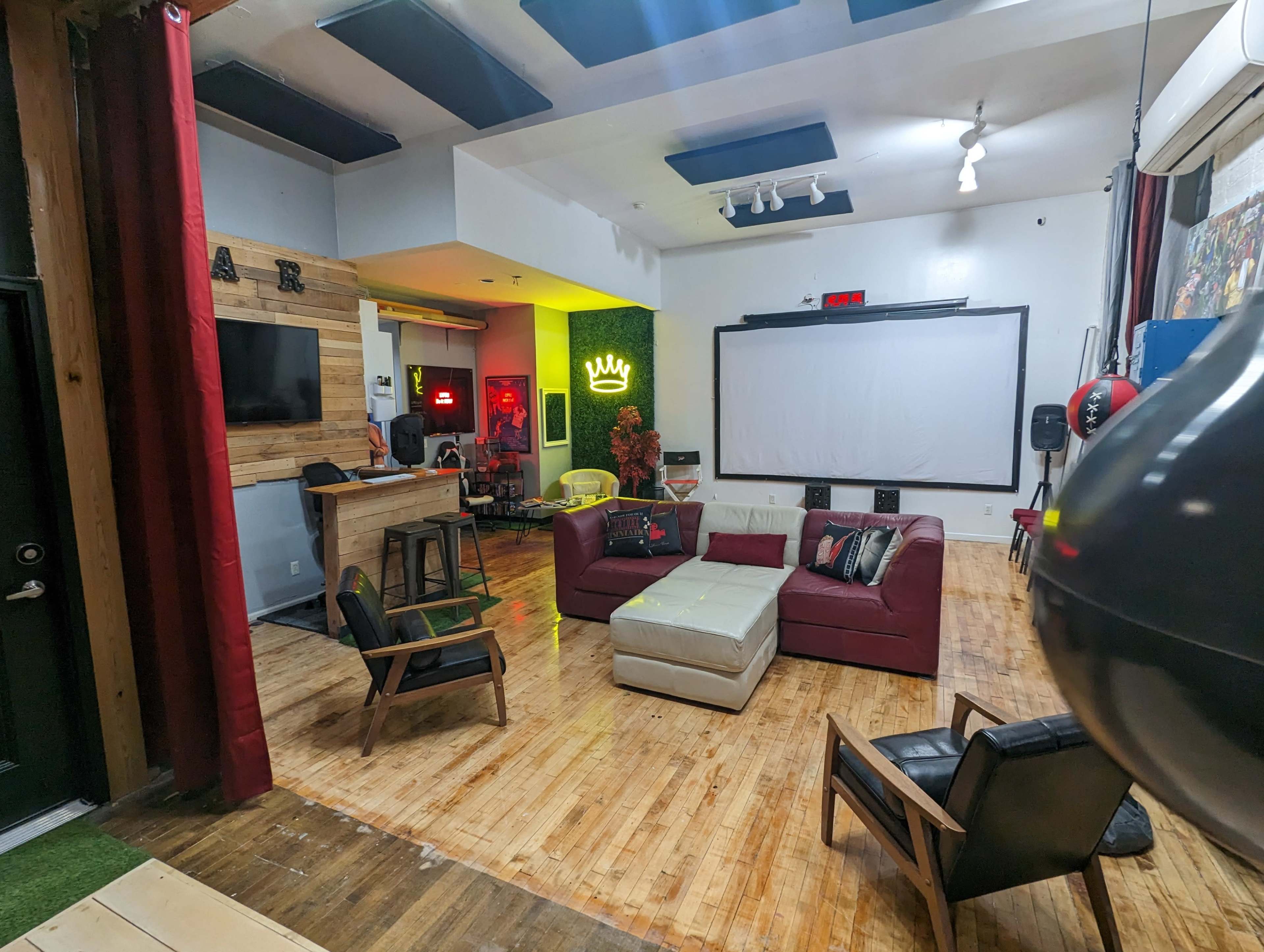 A spacious, modern lounge area with wooden flooring, a central seating arrangement, and a large screen mounted on the wall.