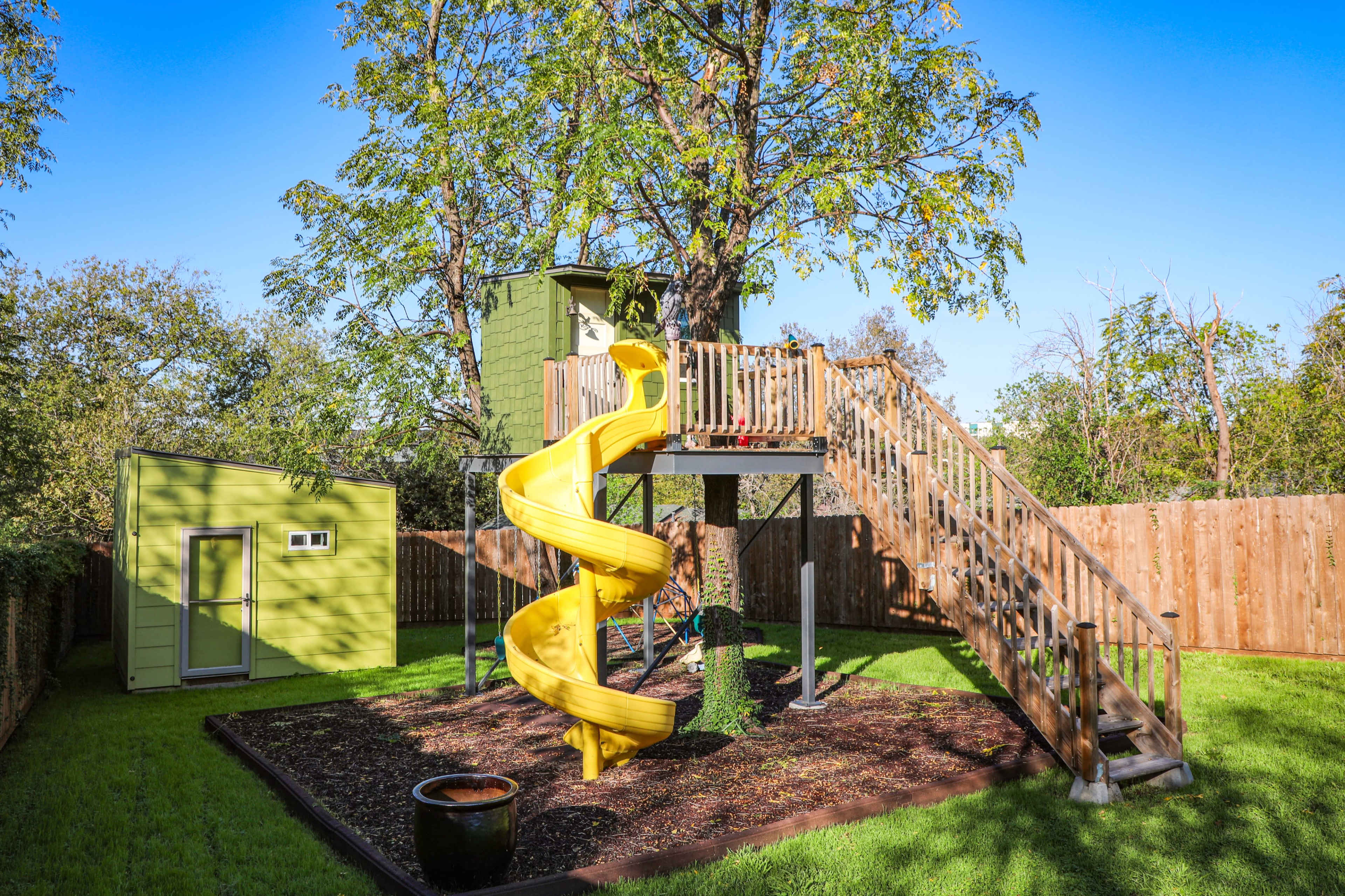 The image shows a backyard playground with a yellow slide, a wooden climbing structure connected to a treehouse, and a small green shed.