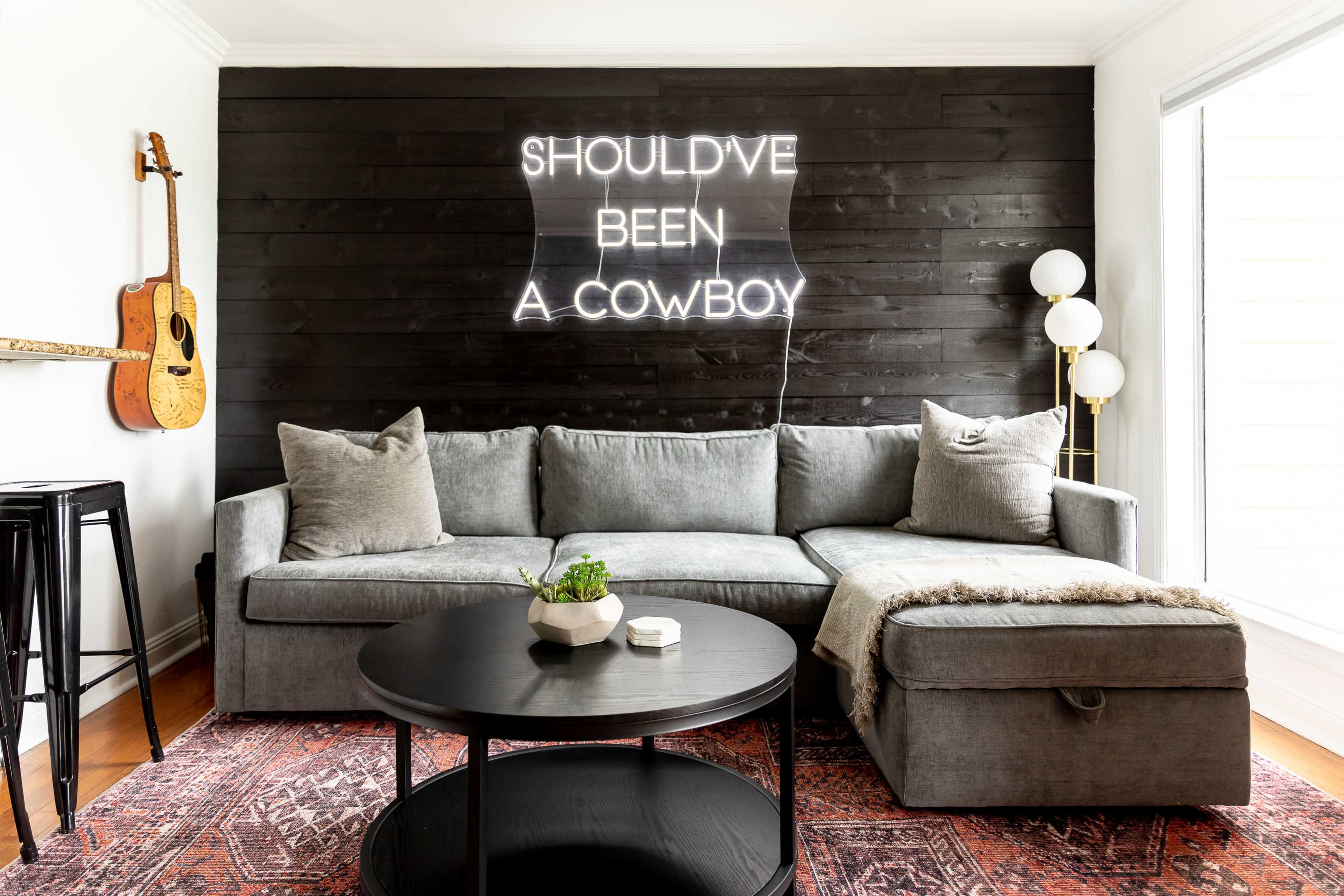 A modern living room features a gray sectional sofa, a round black coffee table, and a neon sign on the wall that reads "SHOULD'VE BEEN A COWBOY," with an acoustic guitar hanging nearby.