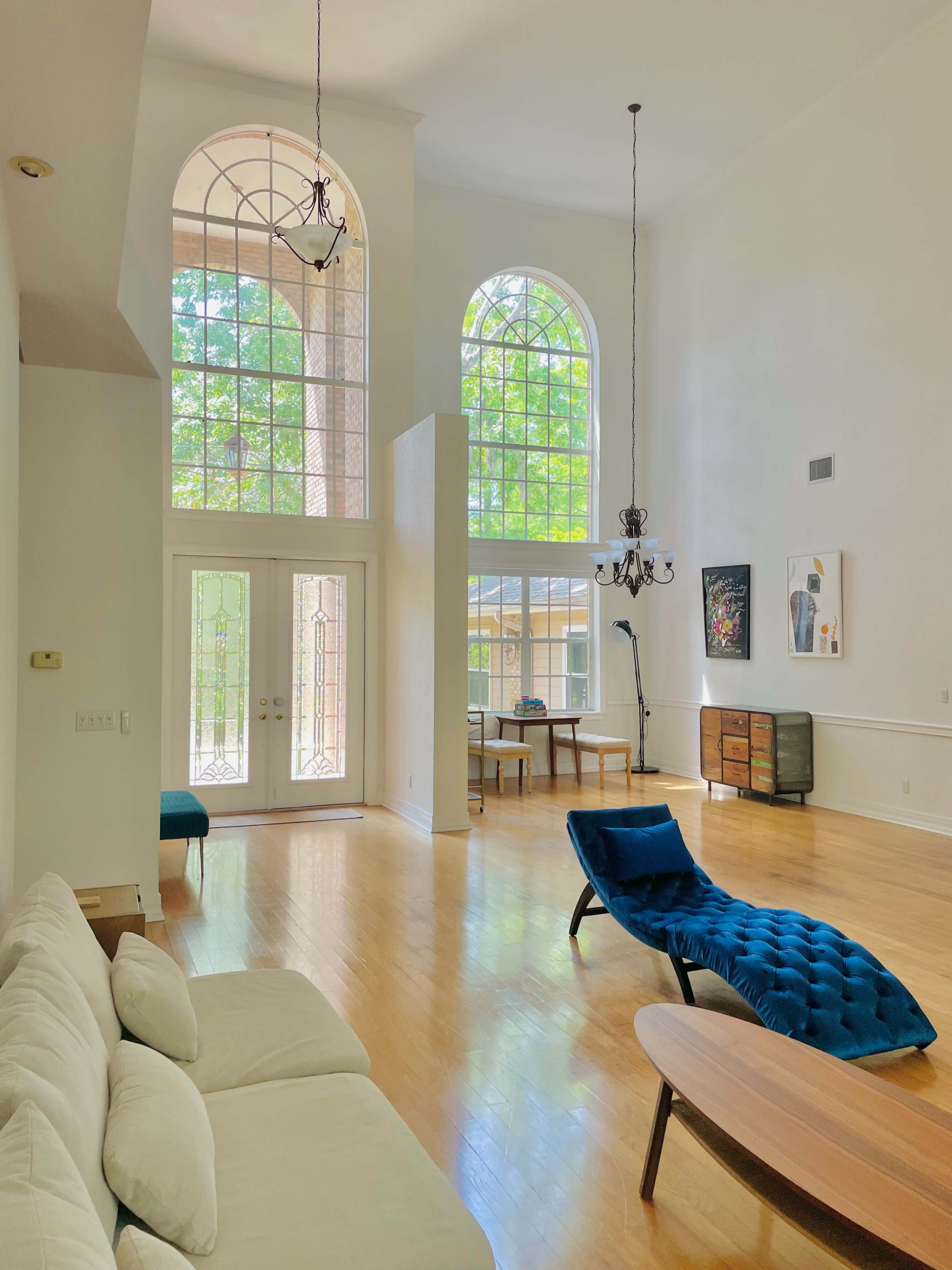 The room features a bright, spacious layout with large arched windows, a light-colored sofa, a blue chaise lounge, a round wooden coffee table, and a chandelier.